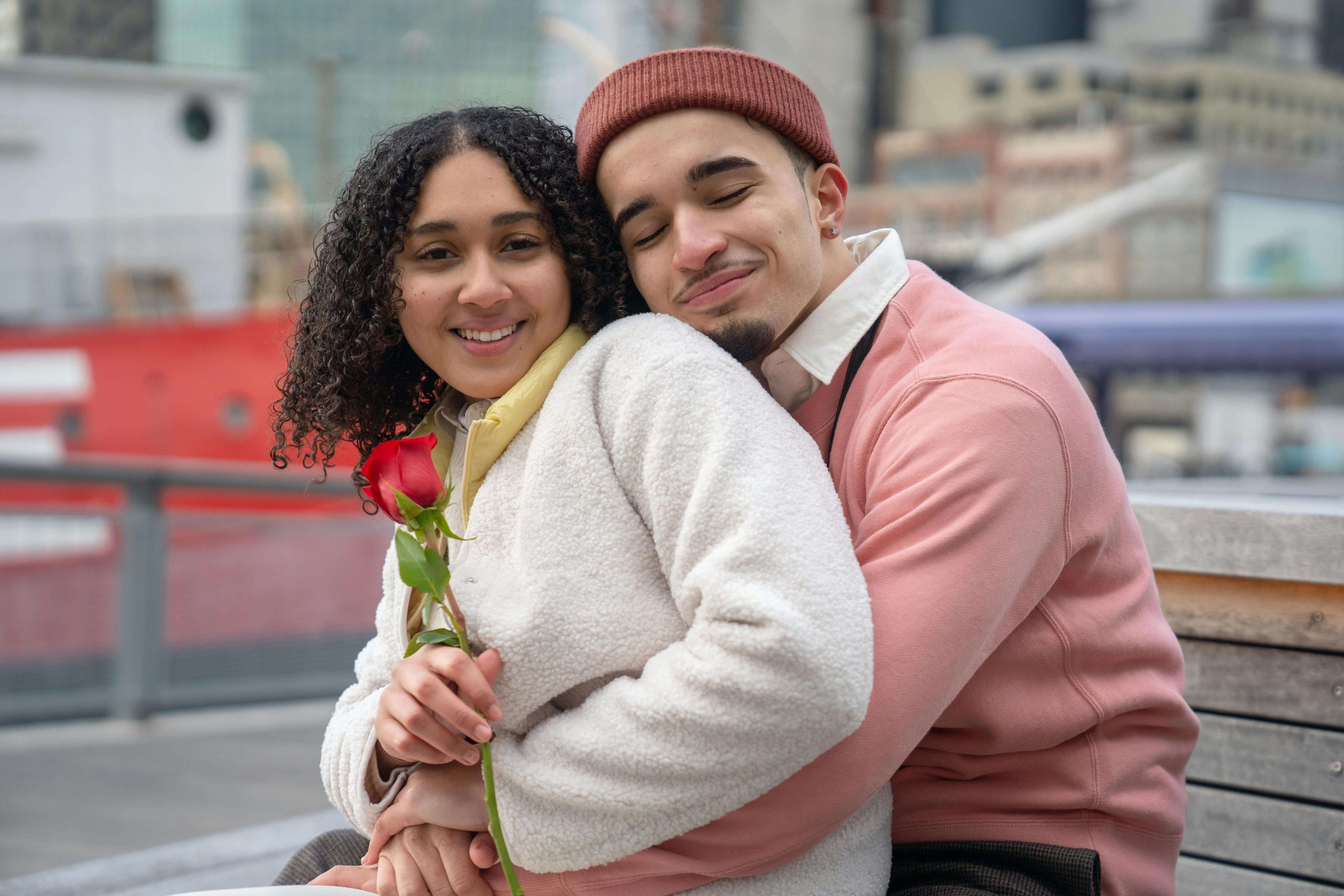 Smiling Latin American couple in warm clothes with red rose cuddling on wooden bench in city street near buildings and ship in daytime