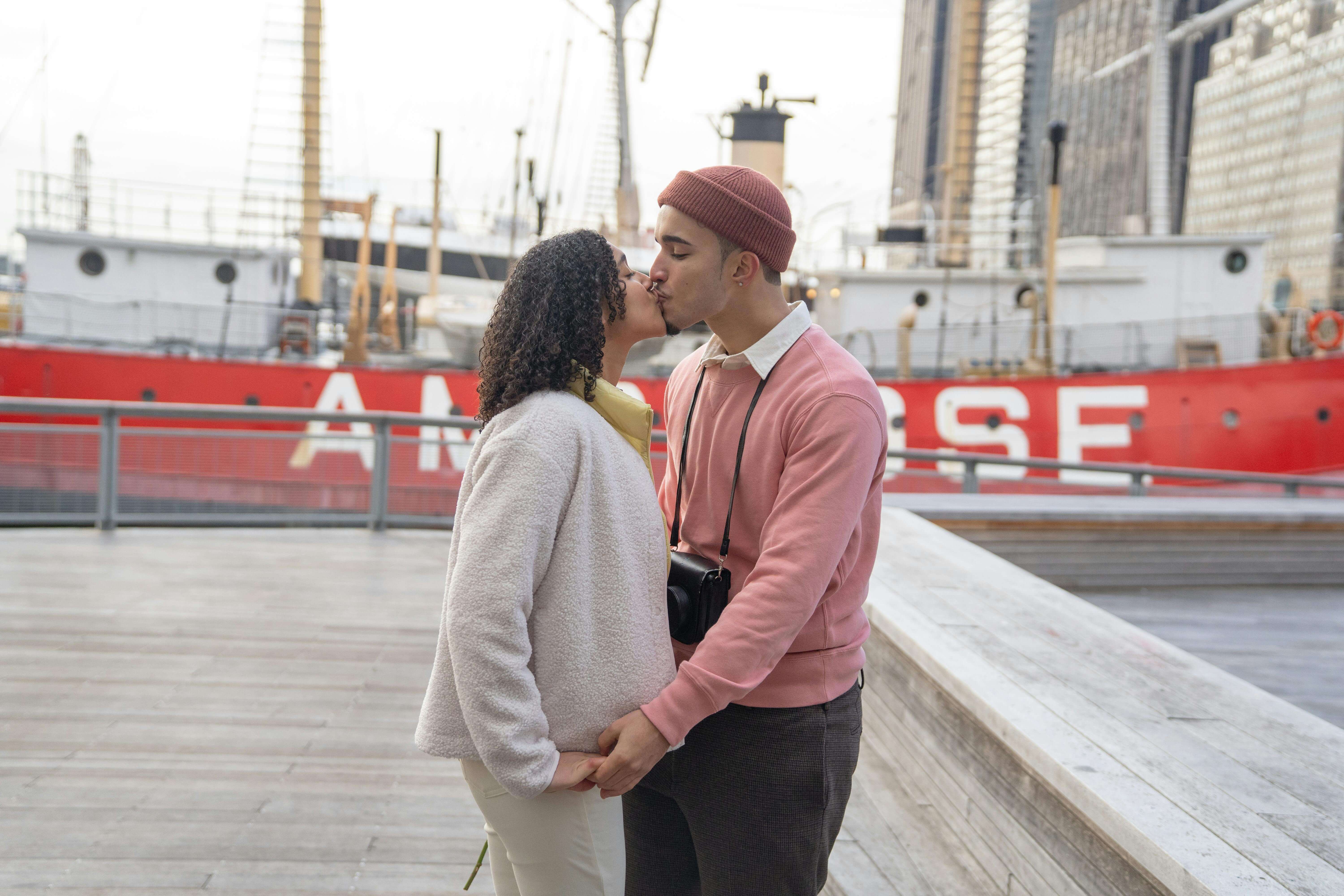 A loving couple shares a kiss by the urban waterfront, capturing a moment of romantic affection.