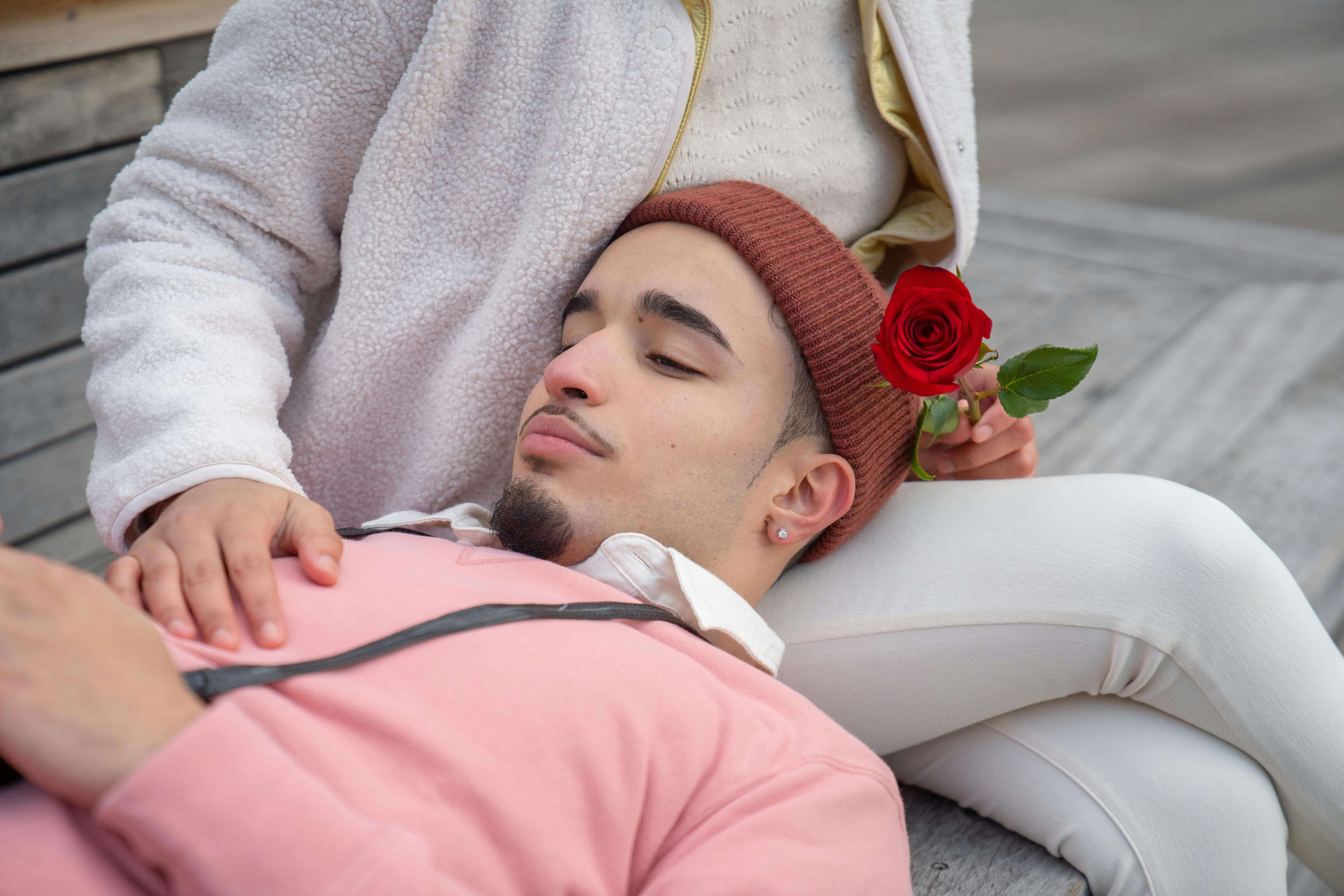 Young Latin American male lying on female laps with red rose on bench in city street in daylight