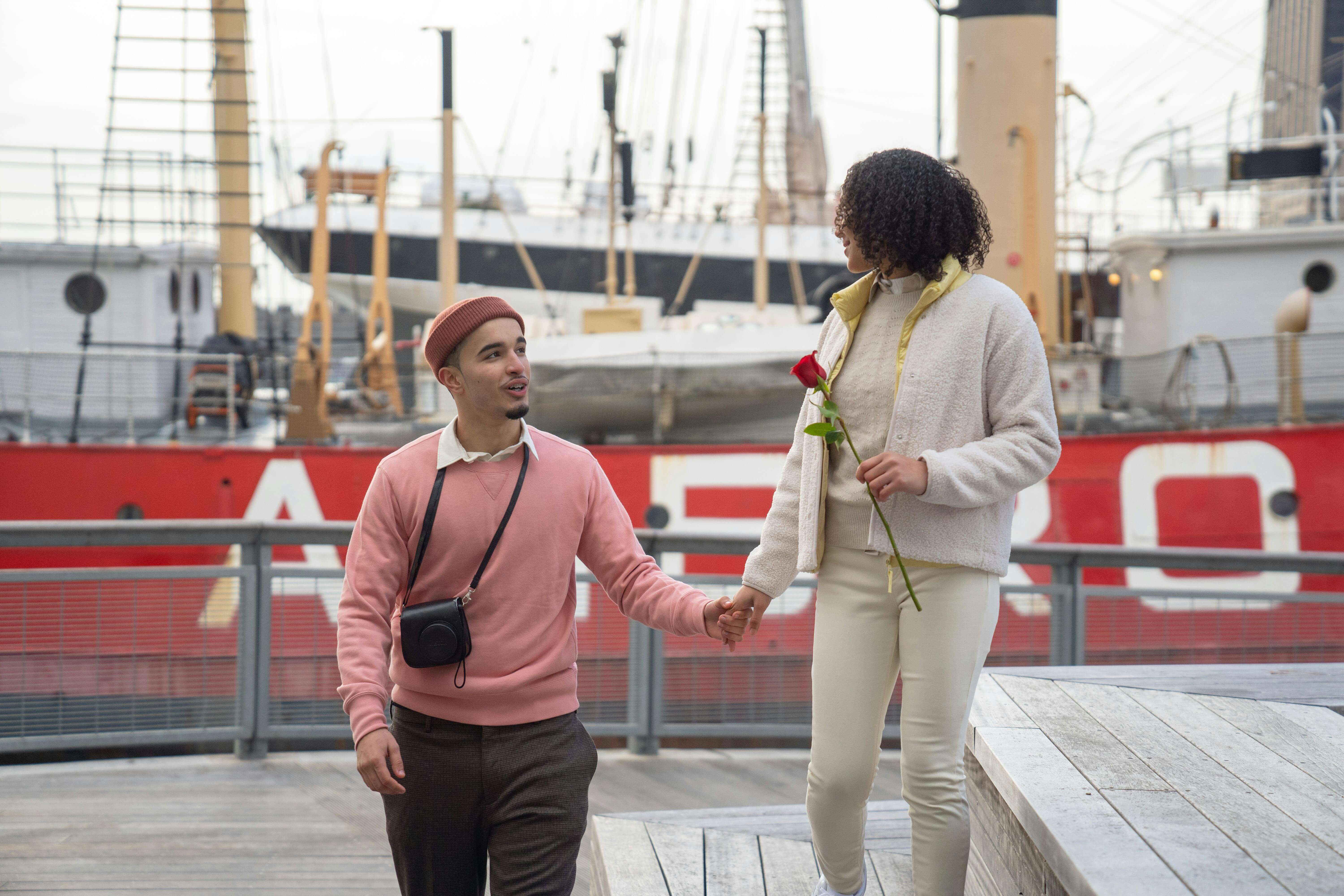 A couple enjoys a romantic walk by the waterfront, holding a rose and hands, against a nautical backdrop.
