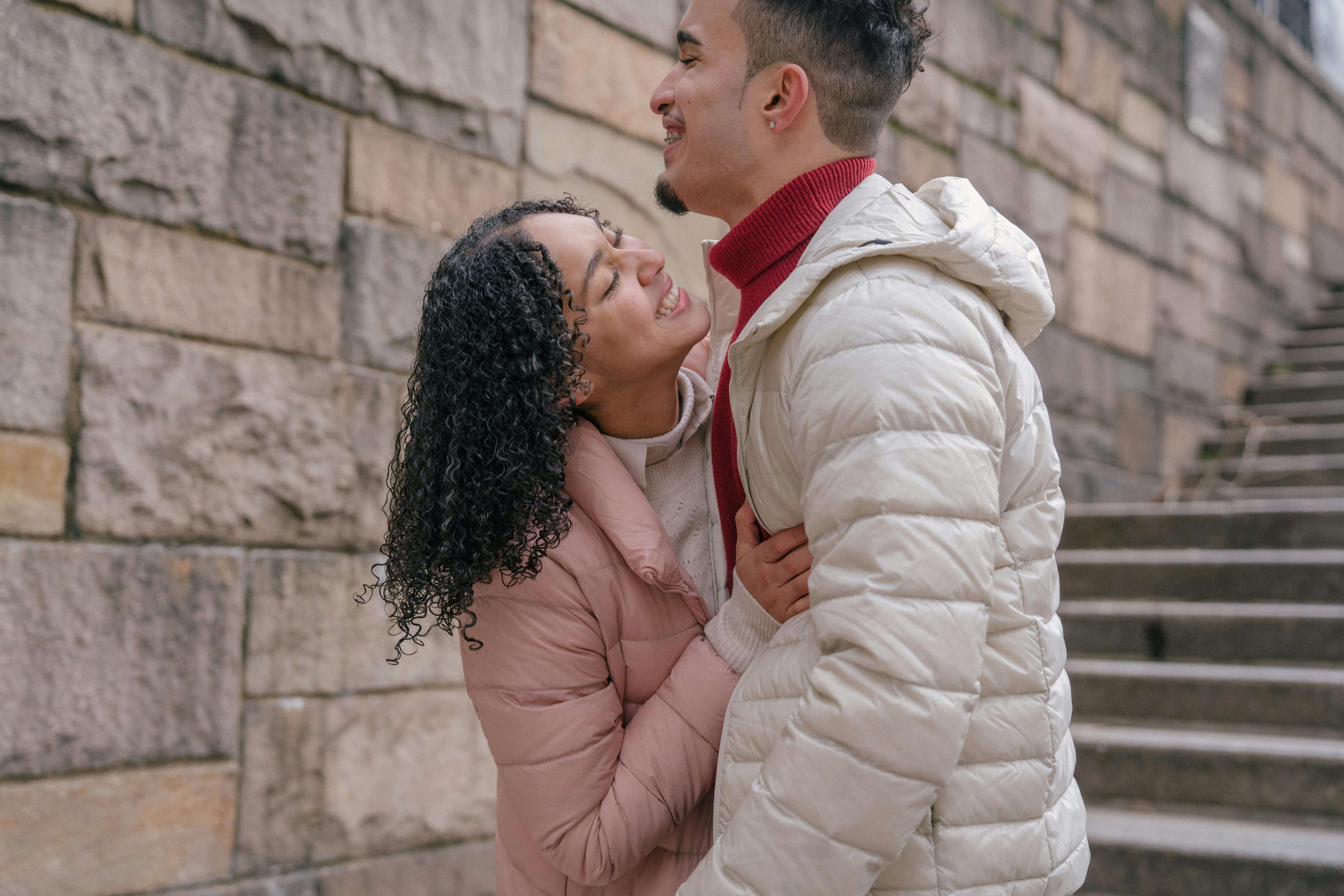 Young couple joyfully embracing on stone steps, wearing warm coats on a city street.