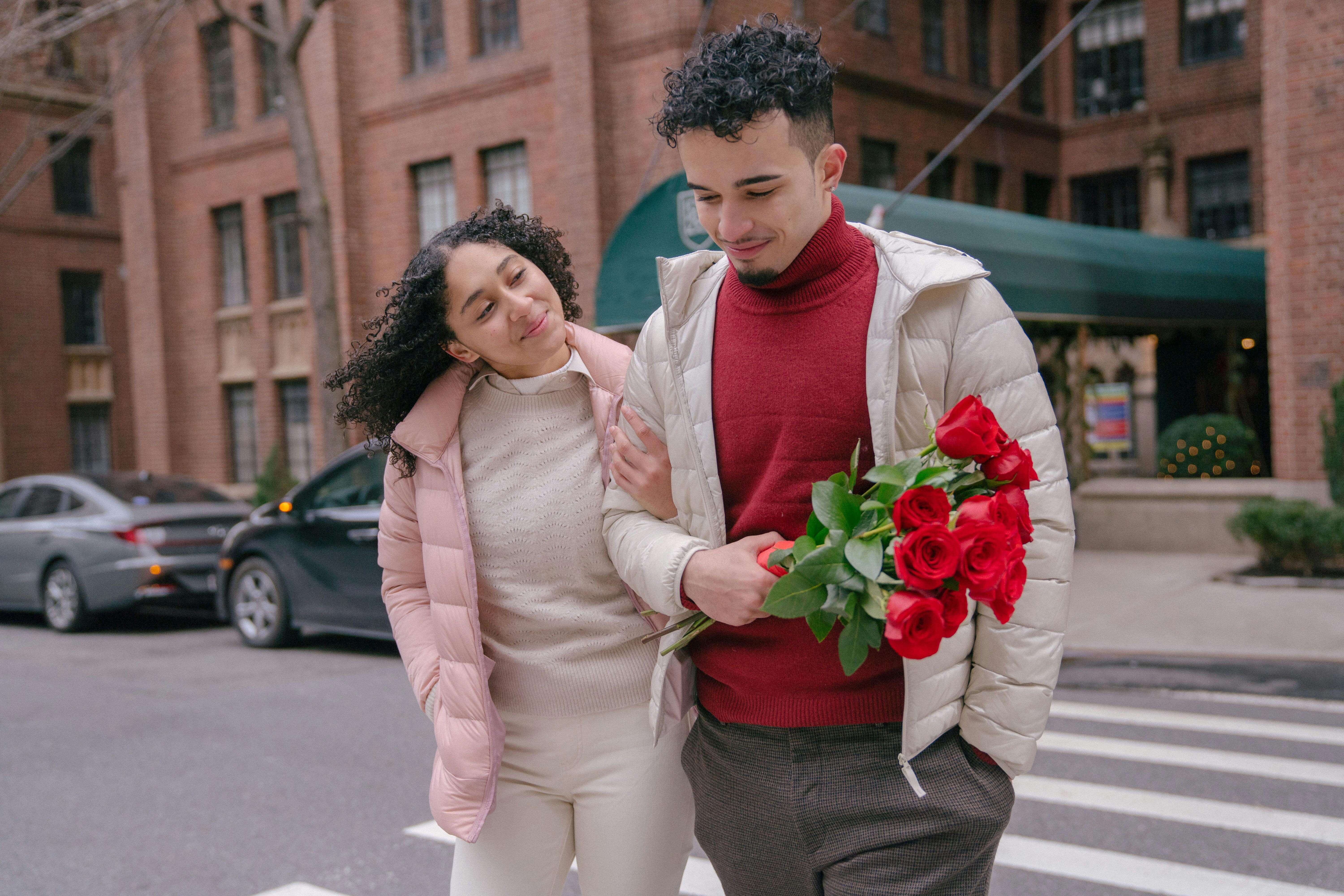 Happy ethnic couple with bright red bouquet of blooming roses crossing road in daytime