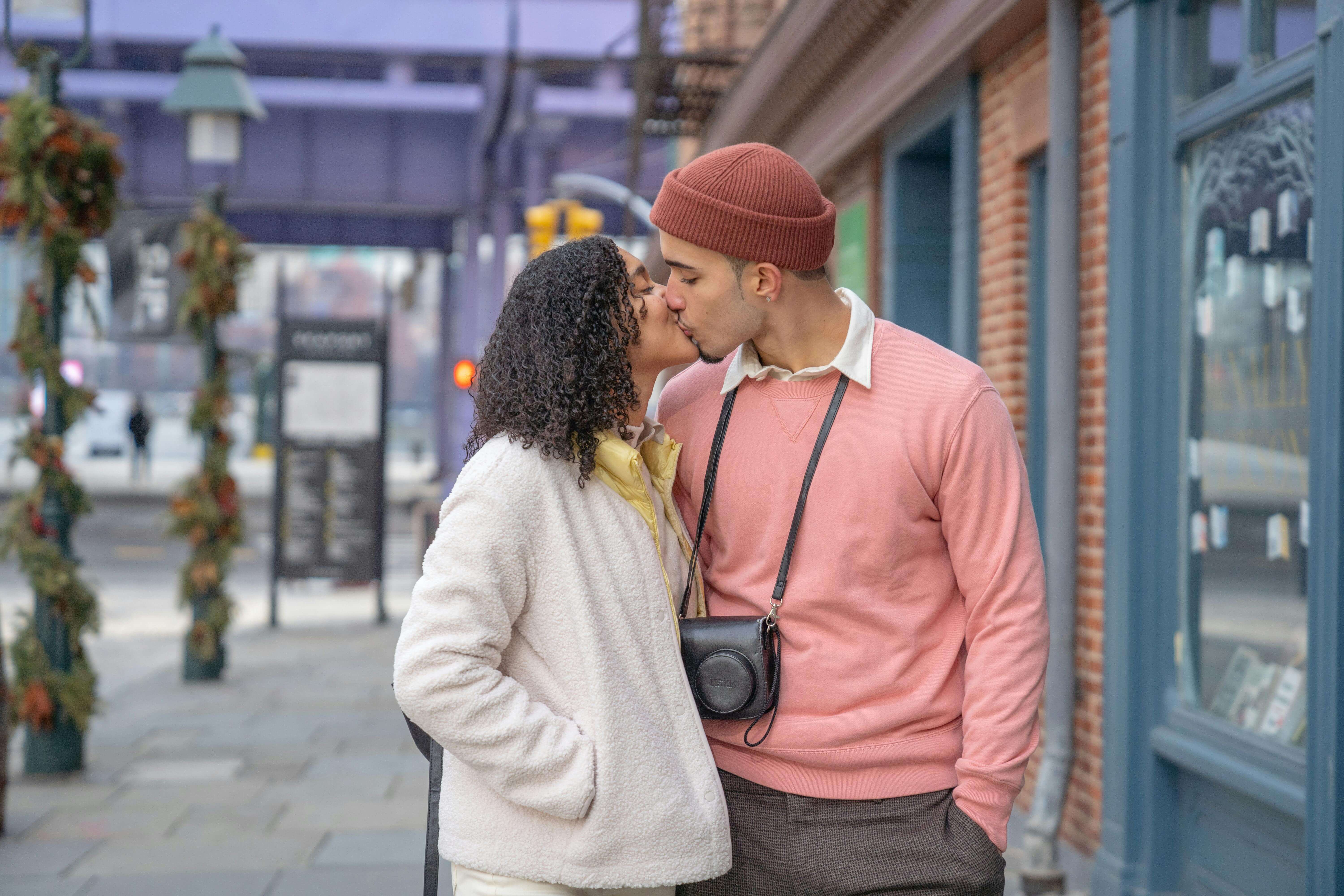 Side view of beloved young ethnic couple in trendy outfits kissing each other standing on city street