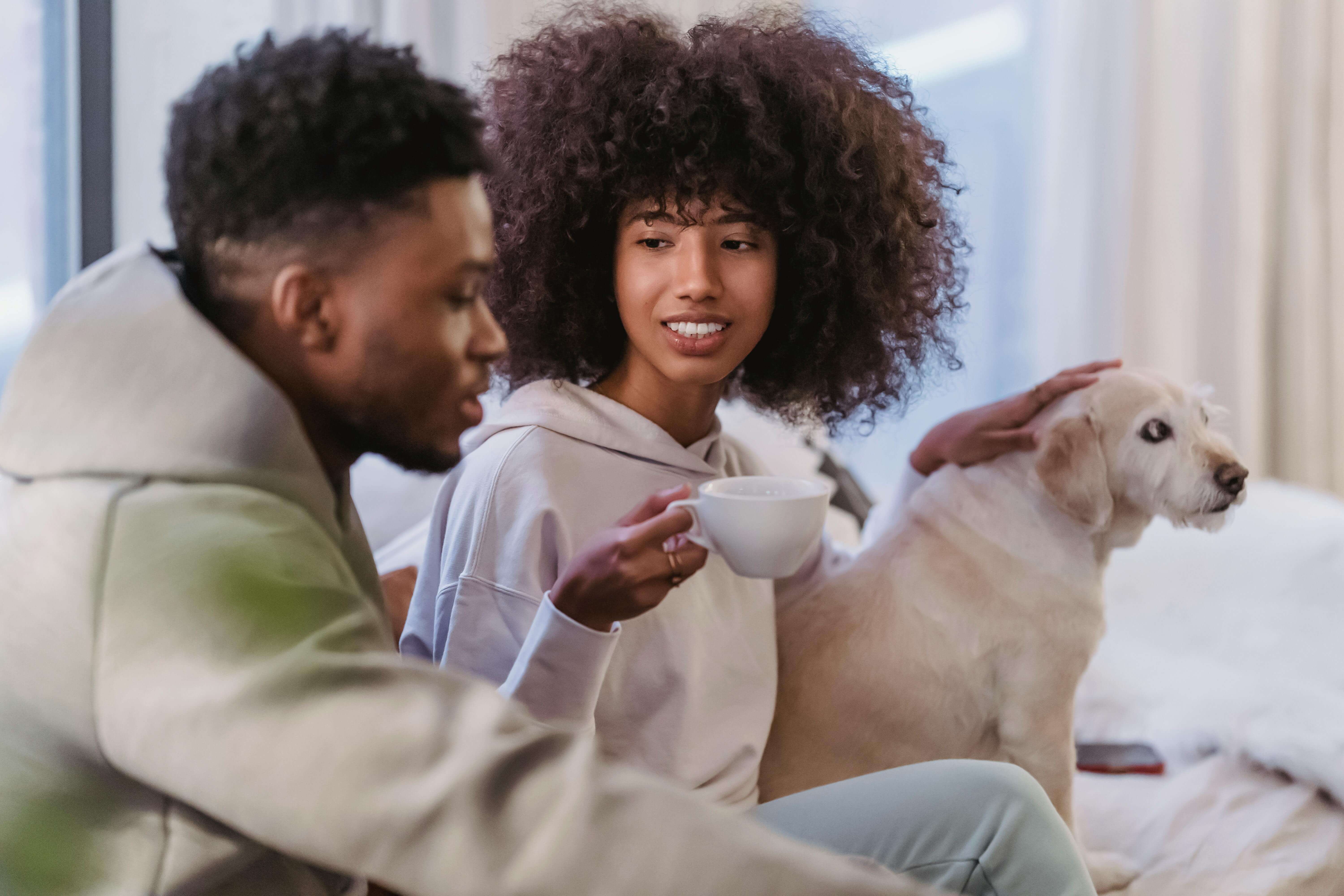 Cheerful African American girlfriend with cup of coffee listening to ethnic boyfriend near funny fluffy dog