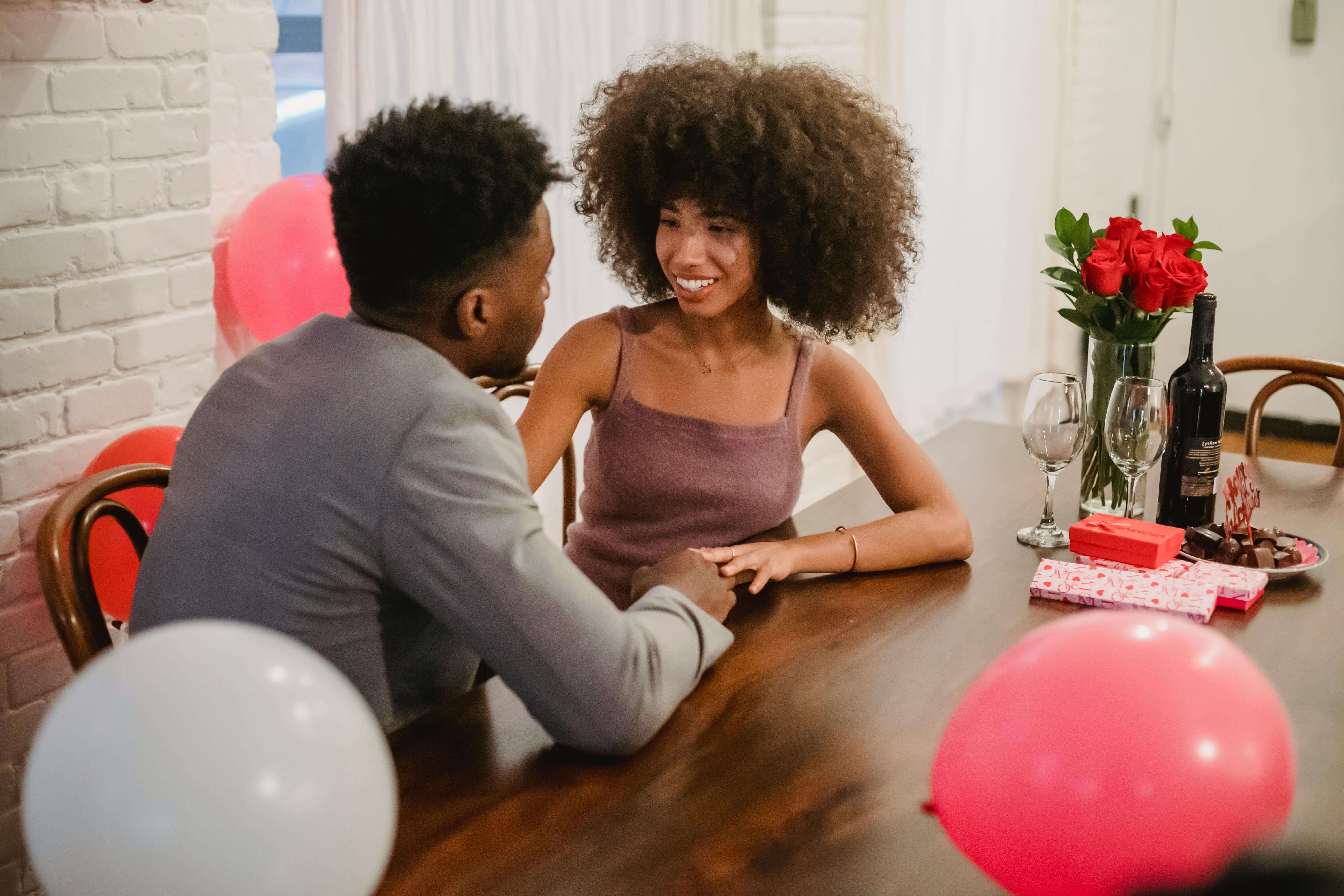 Young black couple in elegant clothes talking while sitting at table with wine bottle and glasses near flowers bouquet in vase and balloons in home while looking at each other