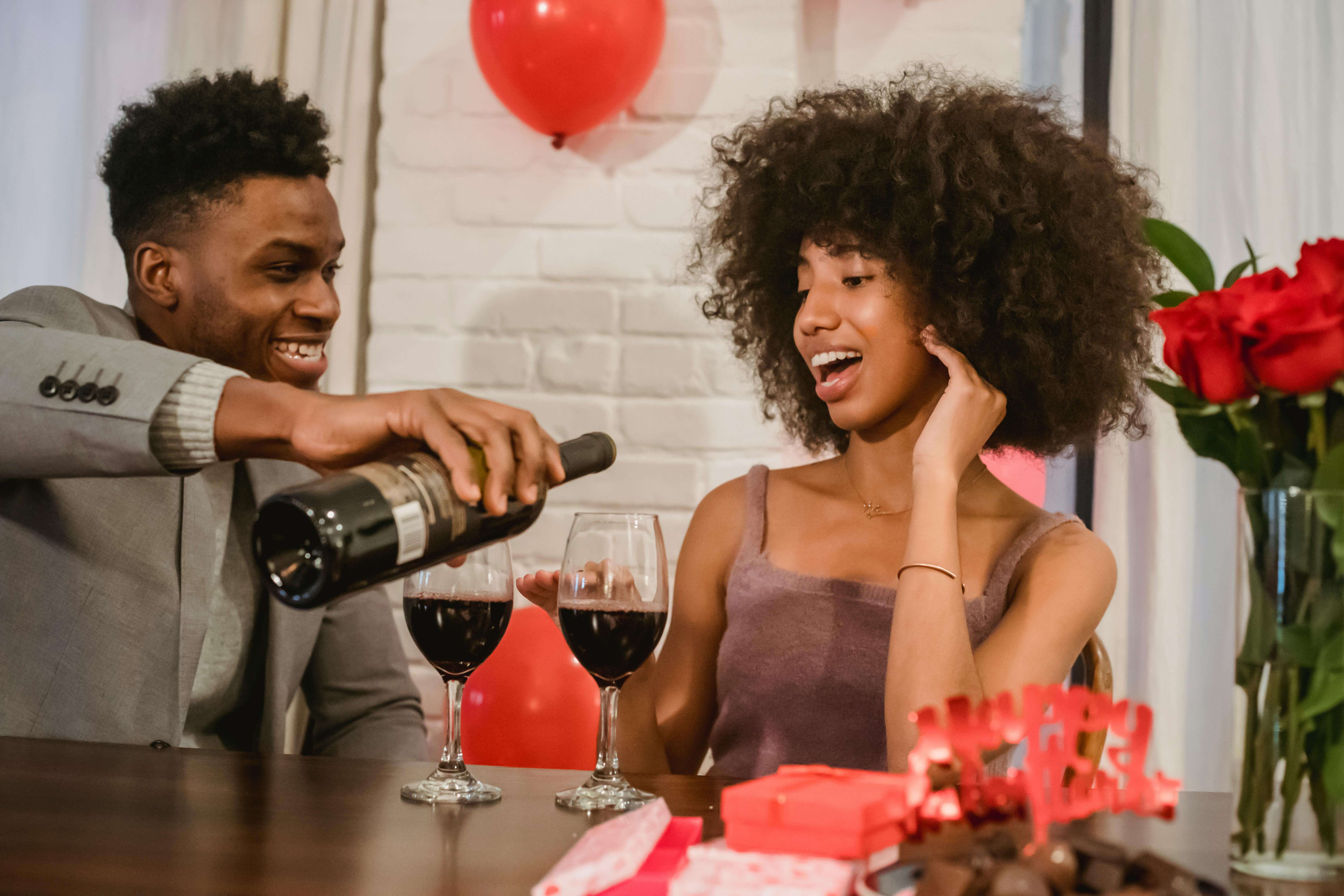 Positive young black couple in elegant clothes sitting at table while man pouring red wine from bottle in glasses near gifts and vase with bouquet with roses at light home