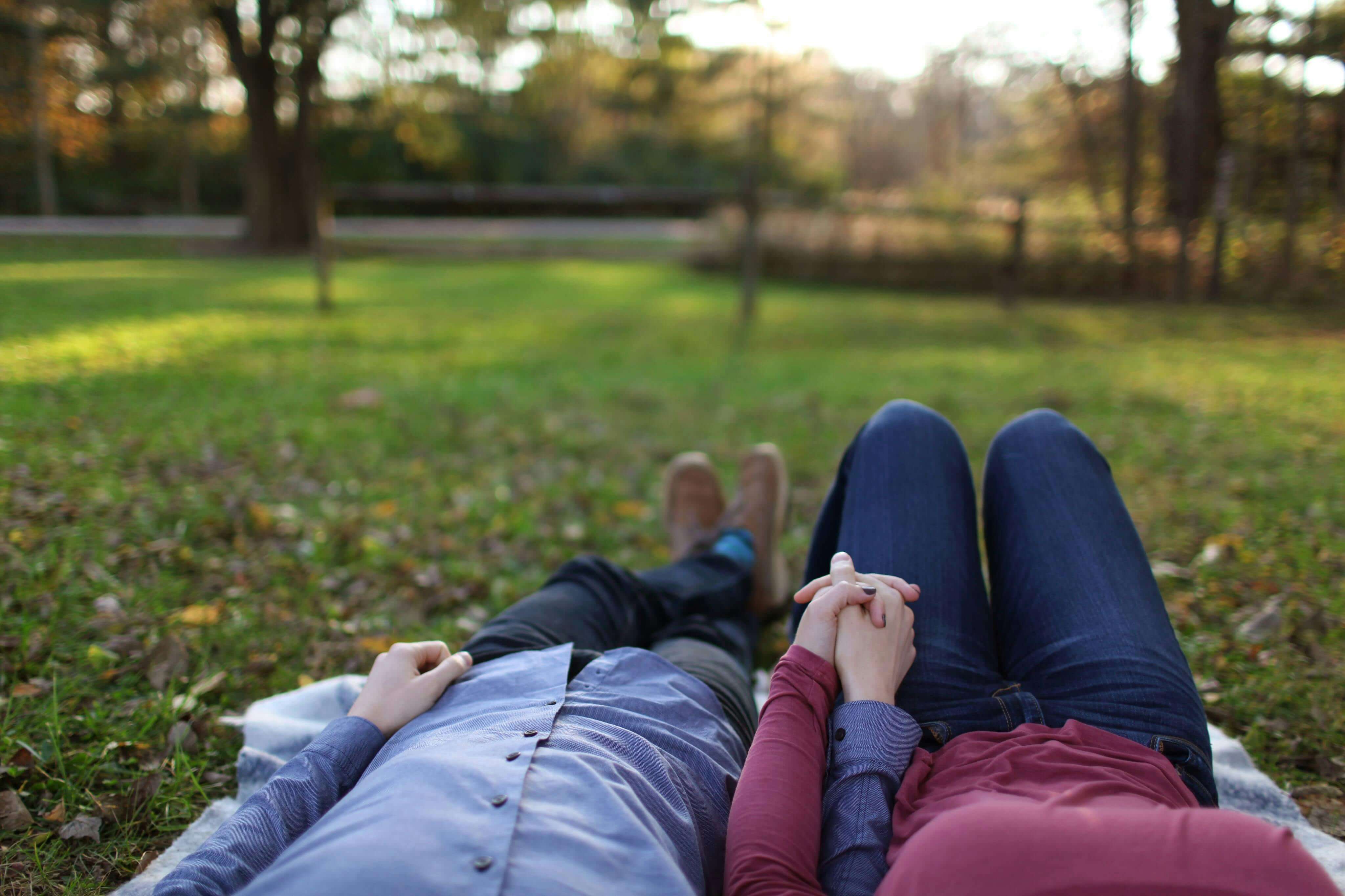 A romantic couple holds hands while lying on grass during a sunny day in the park.
