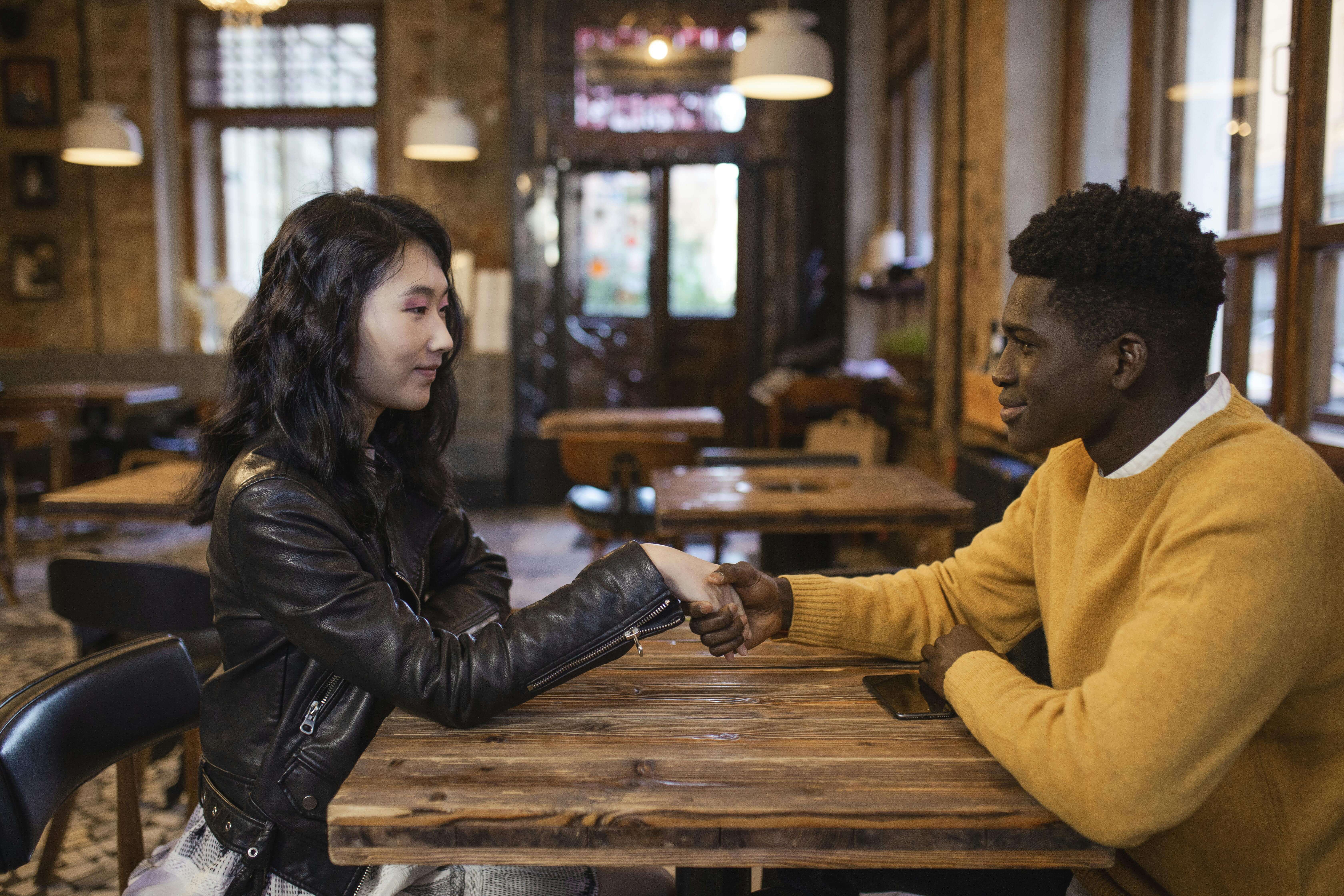 A diverse man and woman sit in a cafÃ©, shaking hands and smiling warmly.