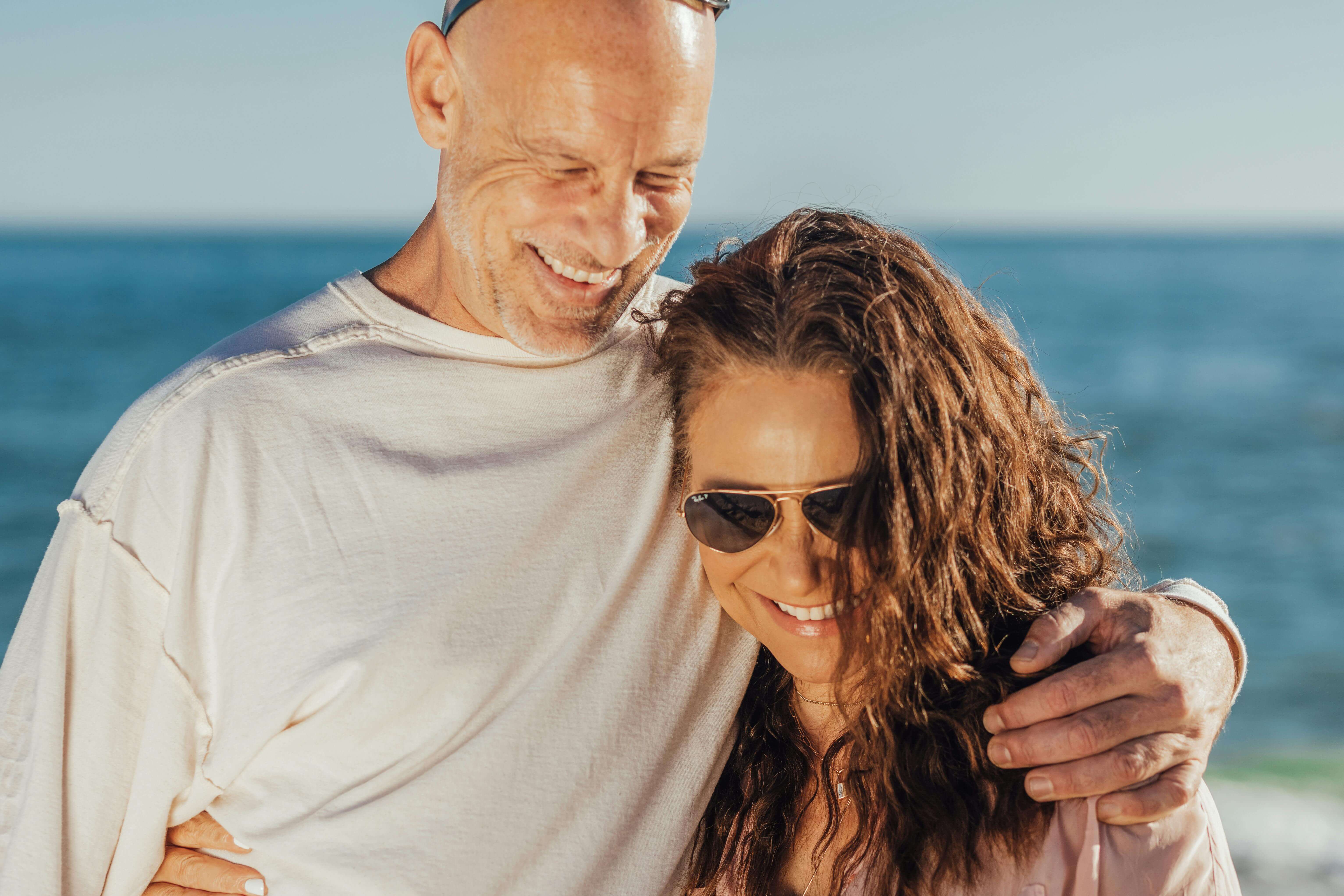 A joyful couple smiling and embracing near the ocean on a sunny day.