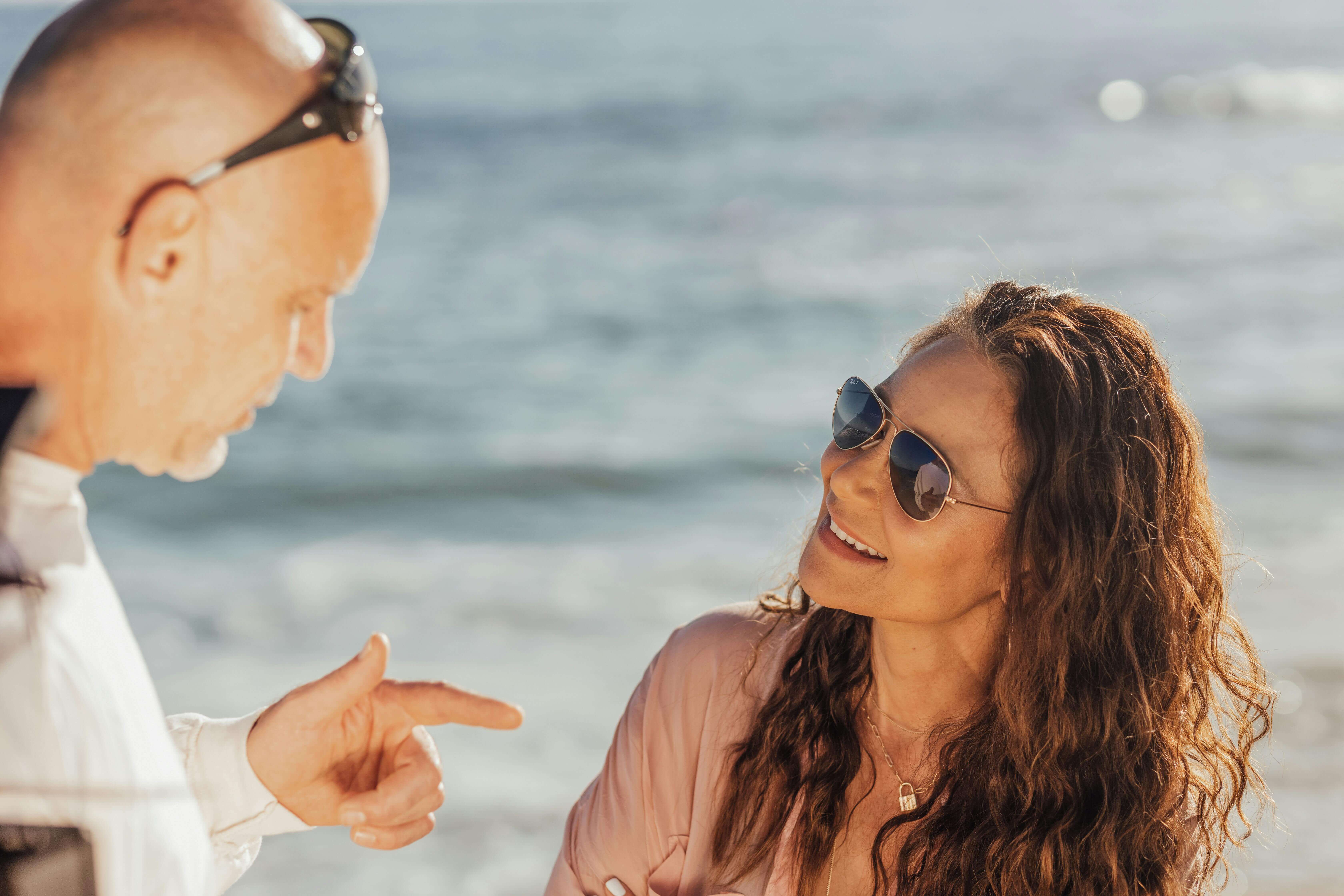 A smiling couple enjoys a casual conversation by the ocean on a sunny day.