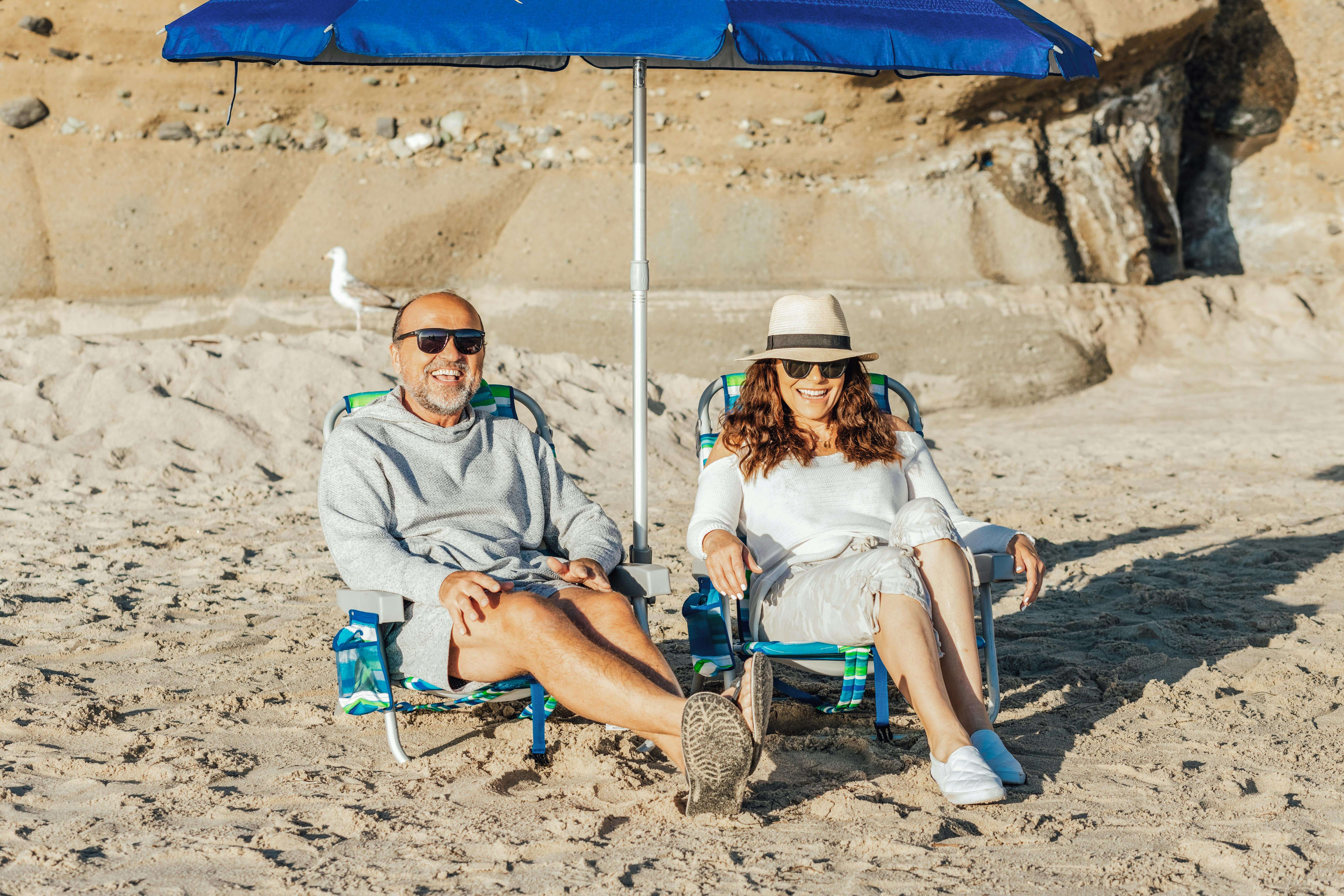 Senior couple enjoys leisurely afternoon on beach, shaded by vibrant blue umbrella.