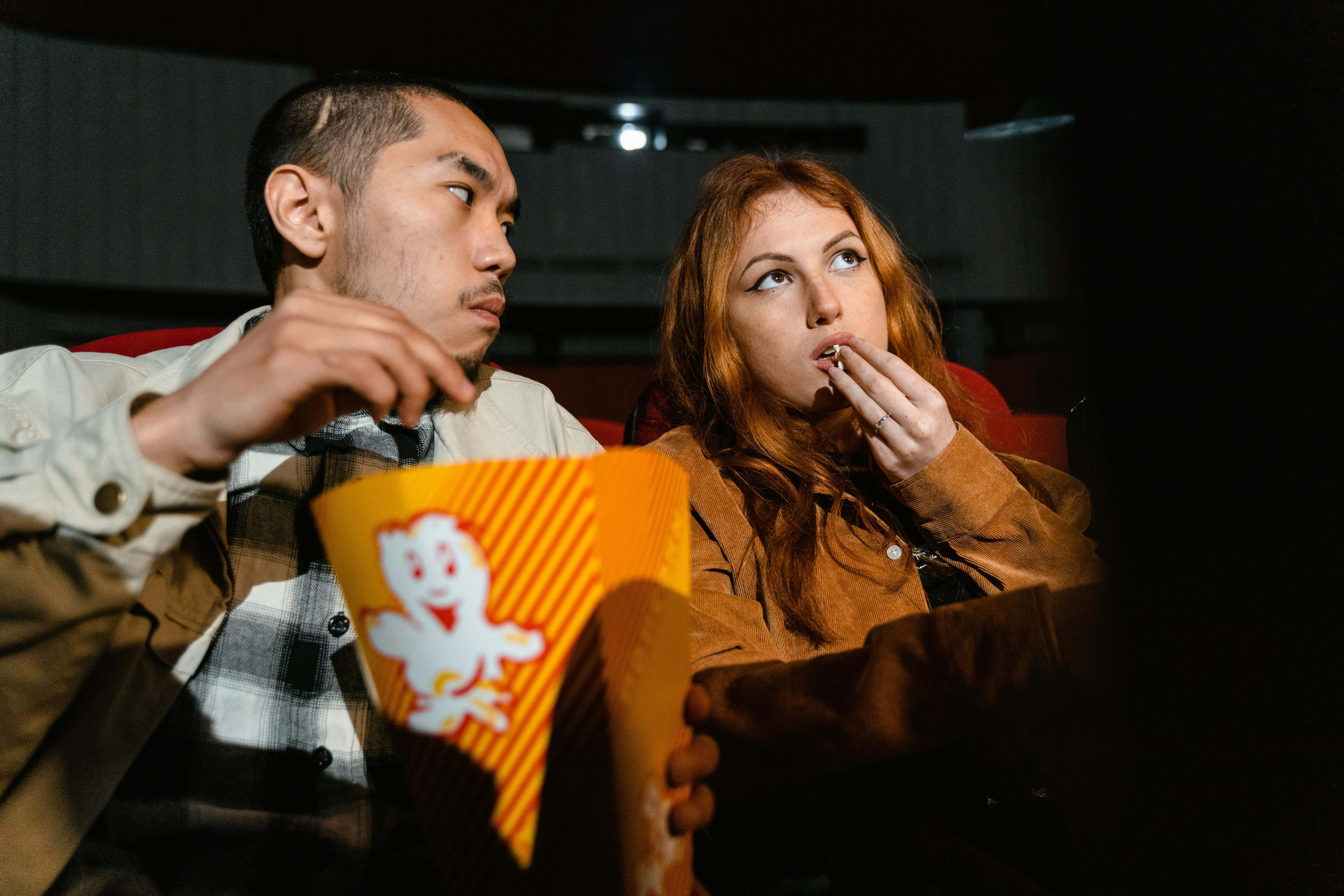 An Asian man and Caucasian woman enjoying popcorn together in a cozy movie theater.