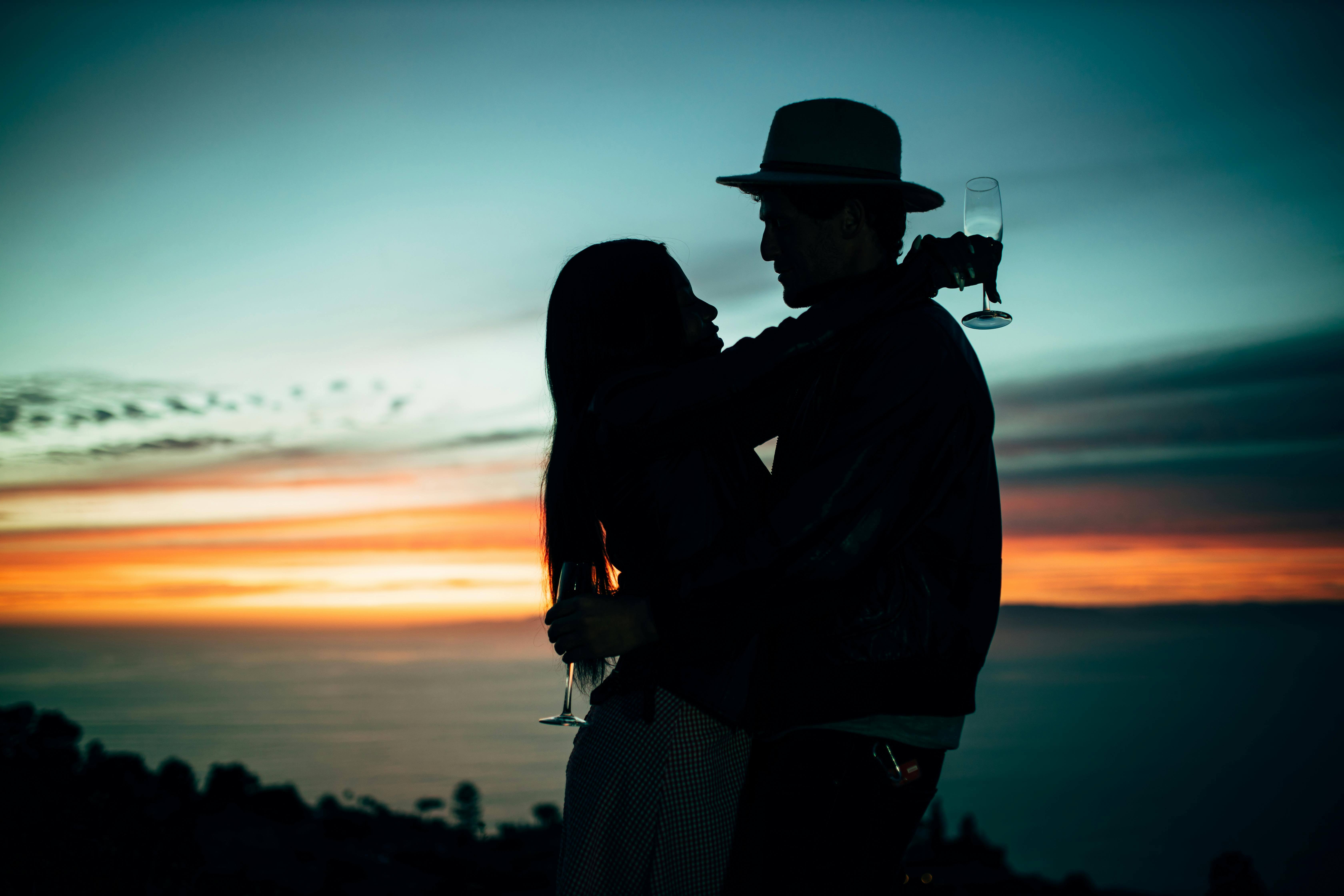 Romantic silhouette of couple embracing with champagne glasses against sunset sky.
