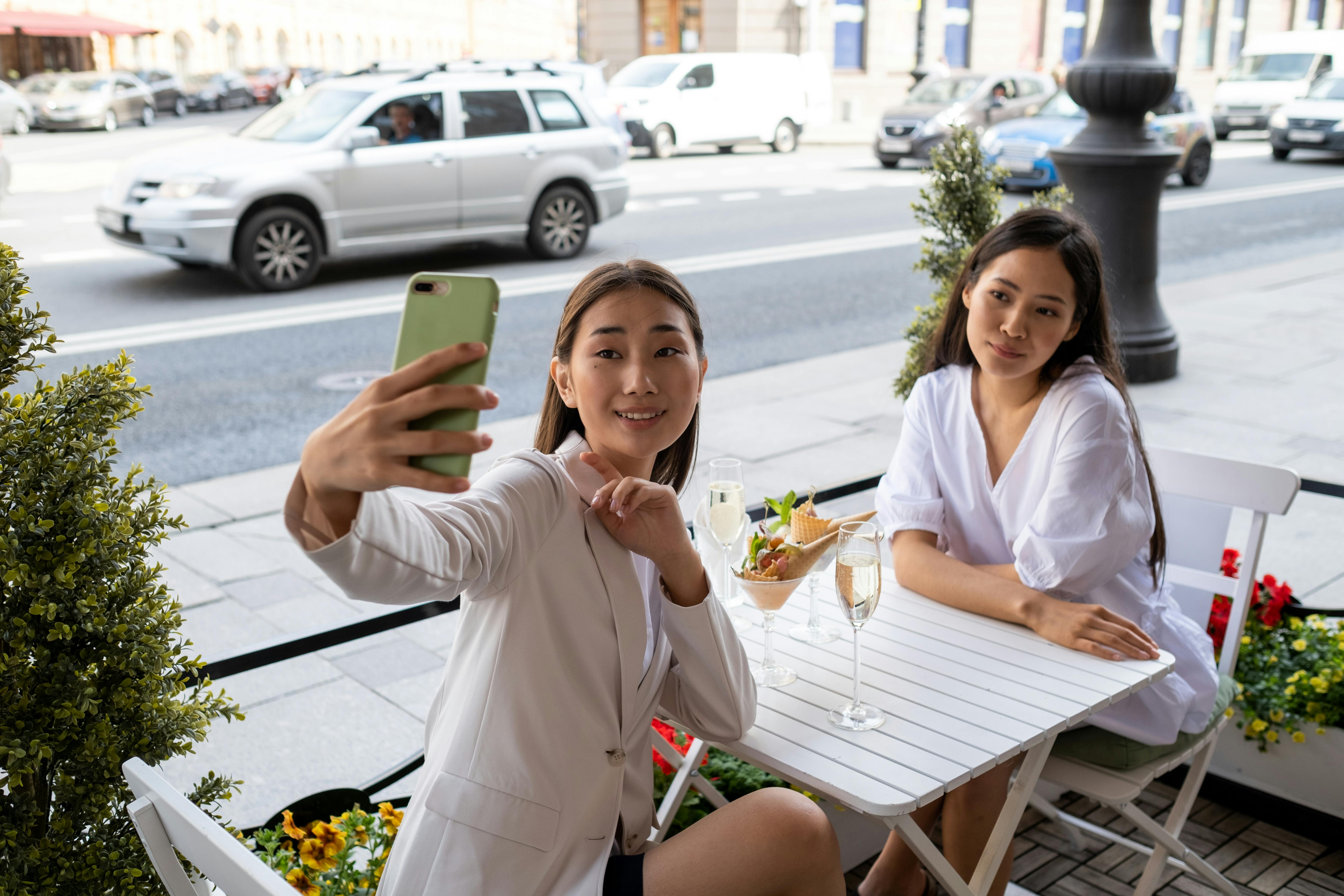 Two women taking a selfie at an outdoor cafe table on a sunny day.