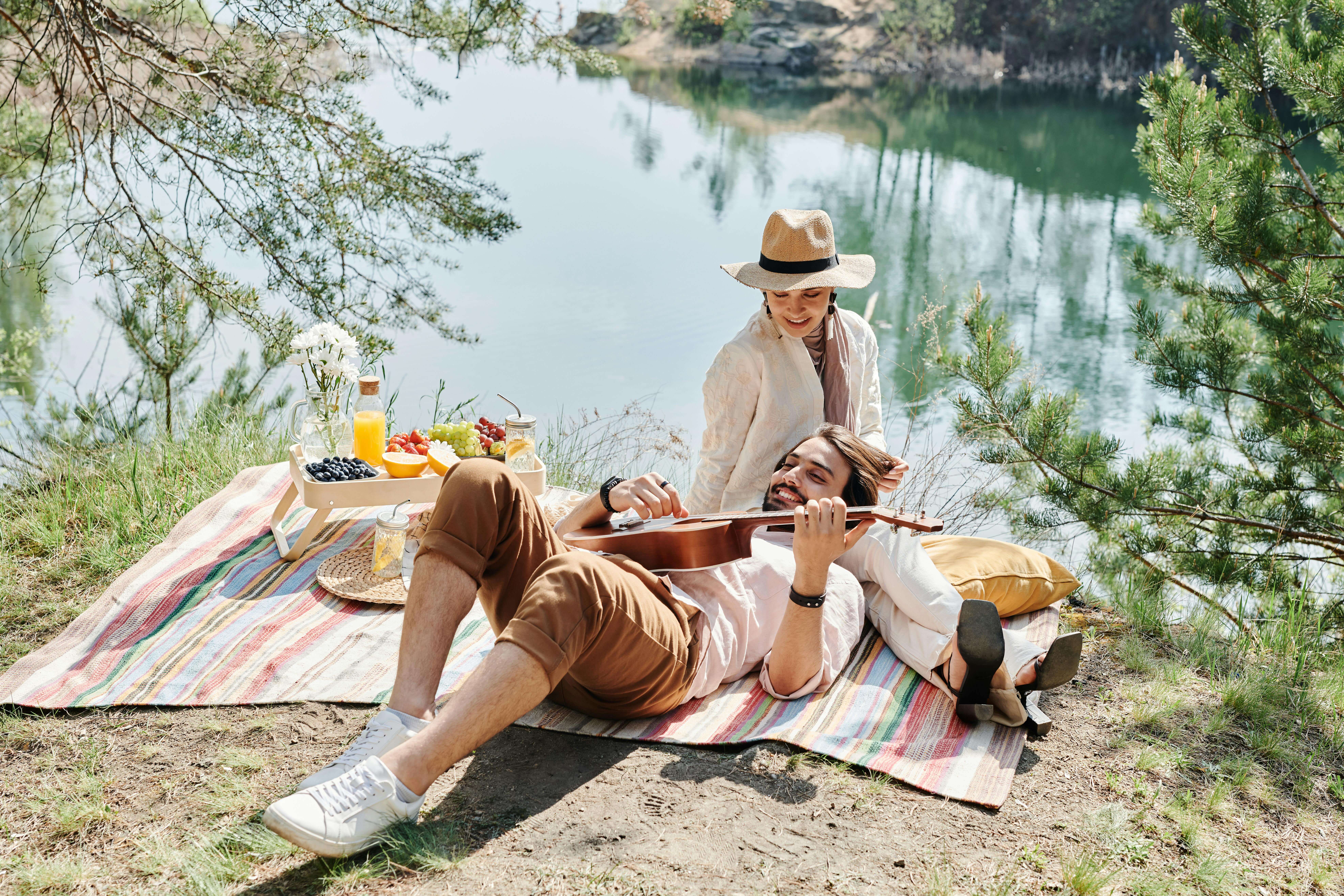 A couple enjoying a serene picnic by the lake, playing ukulele and sharing a joyful moment.