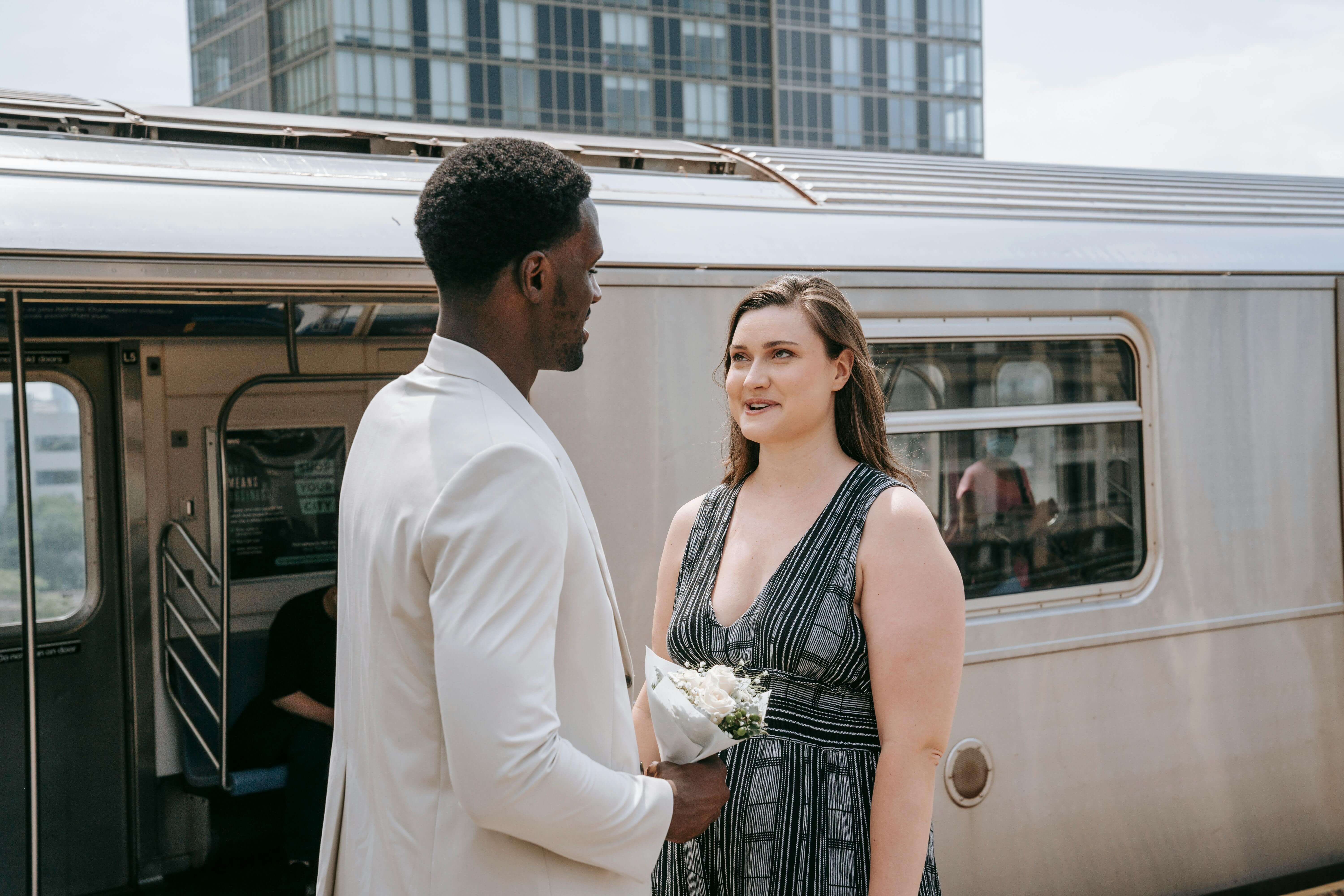A couple stands talking at a train station, holding flowers, with a modern city backdrop.