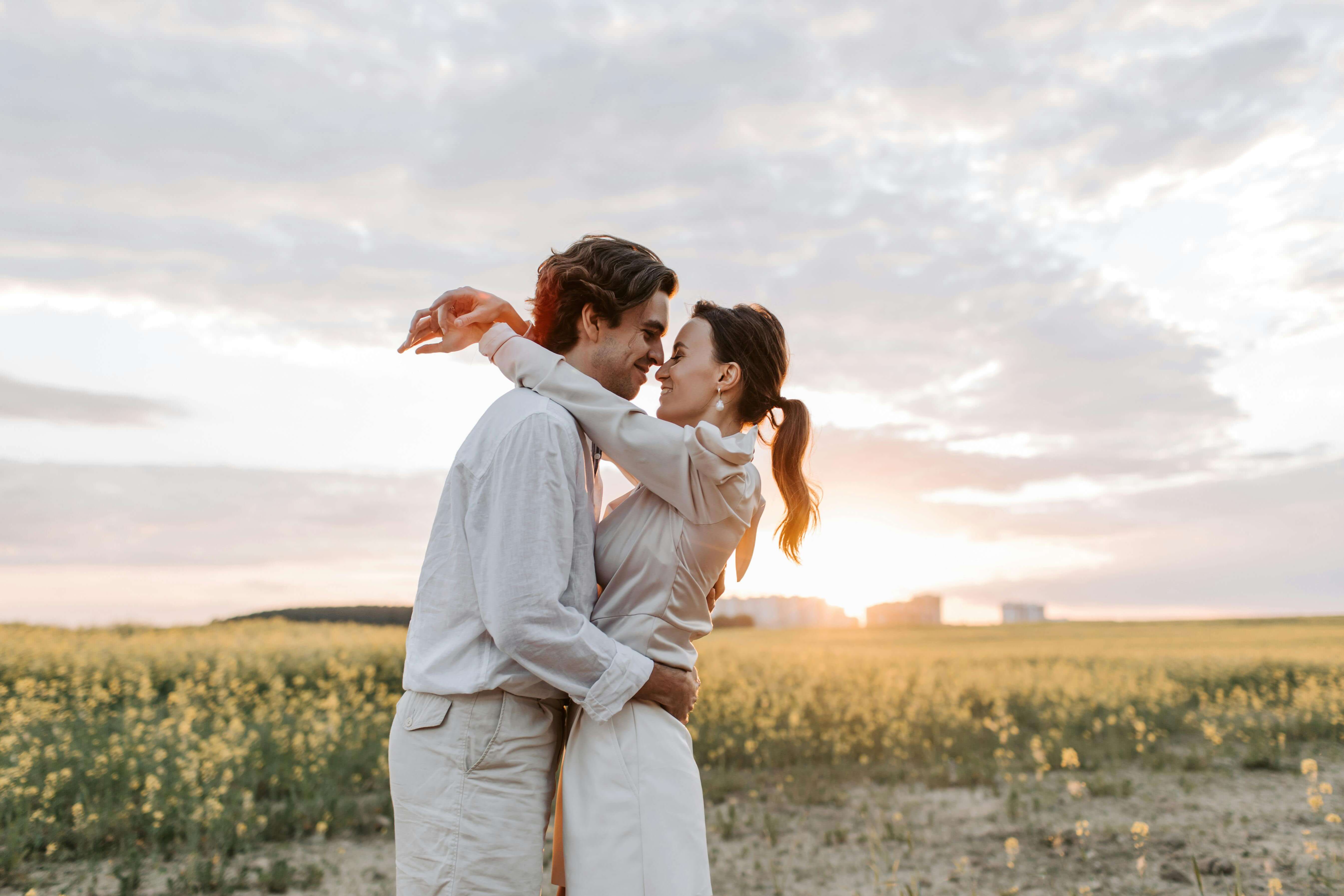 A romantic couple enjoys a tender embrace in a sunny flower field under a serene sky.