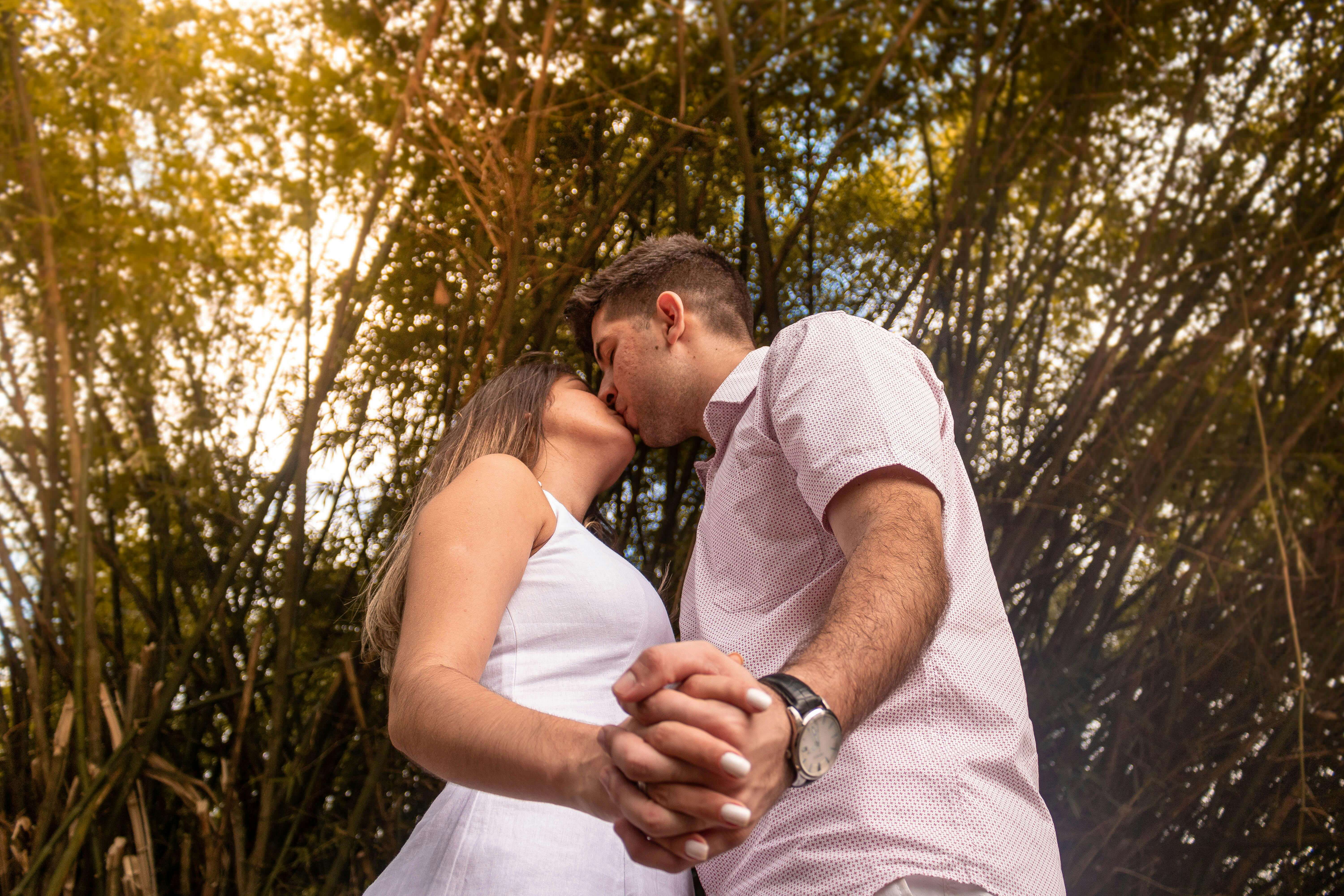 A couple sharing a romantic kiss while holding hands in a lush outdoor setting.