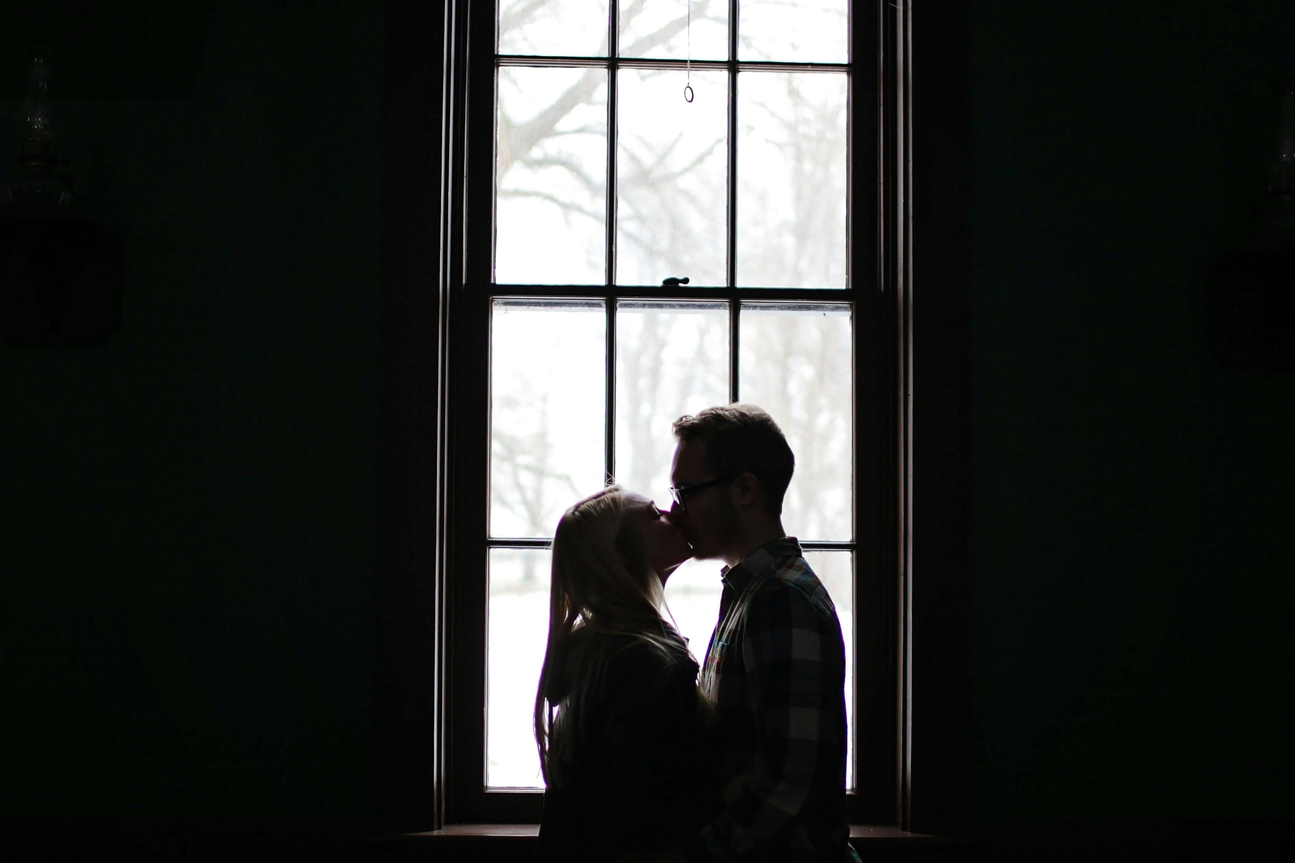 A romantic silhouette of a couple sharing a kiss in front of a large, backlit window.
