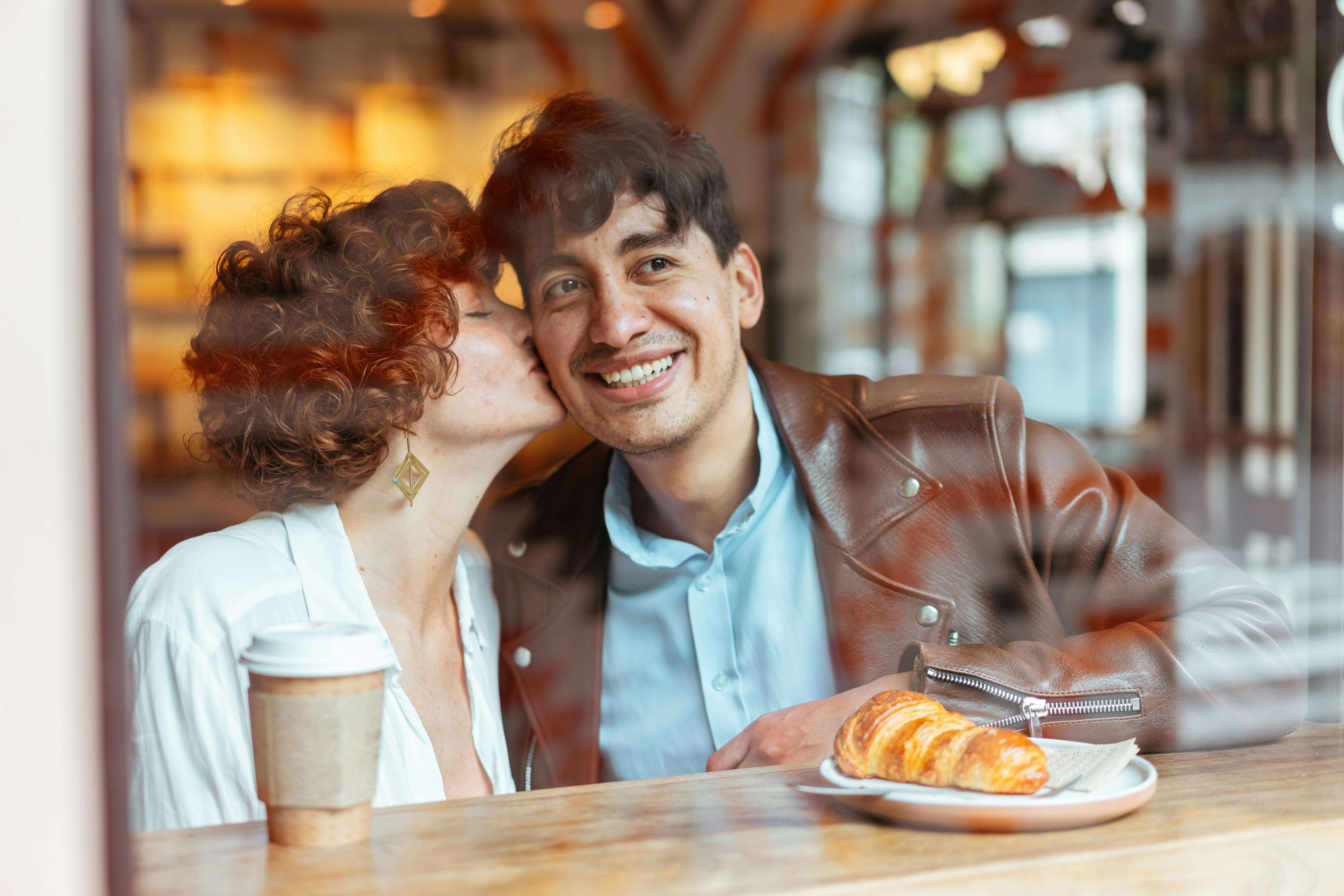 A joyful couple sharing a sweet moment in a cozy cafÃ© setting, enjoying a croissant and coffee.