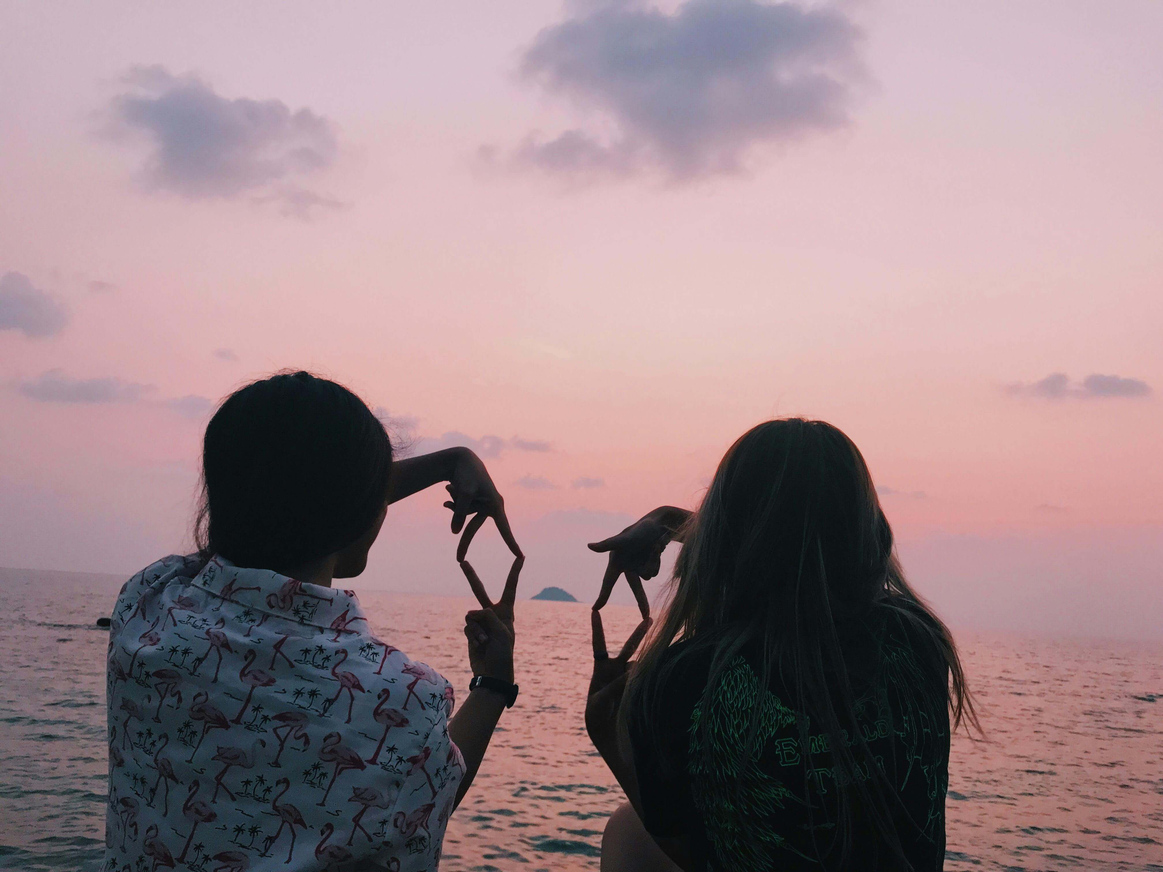 Two friends form heart shapes with their hands by the ocean at sunset, enjoying a peaceful moment.
