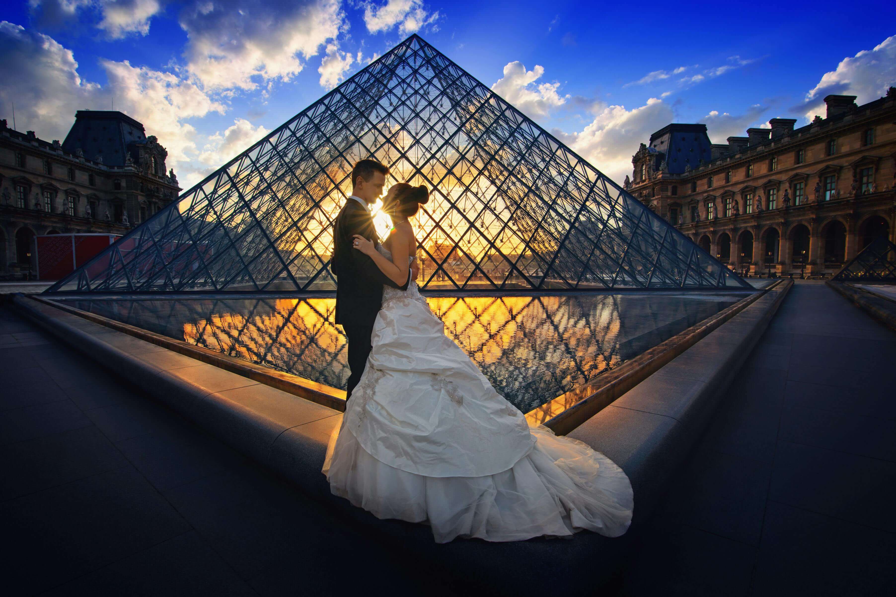Couple embraces before the Louvre Pyramid during a stunning sunset in Paris.