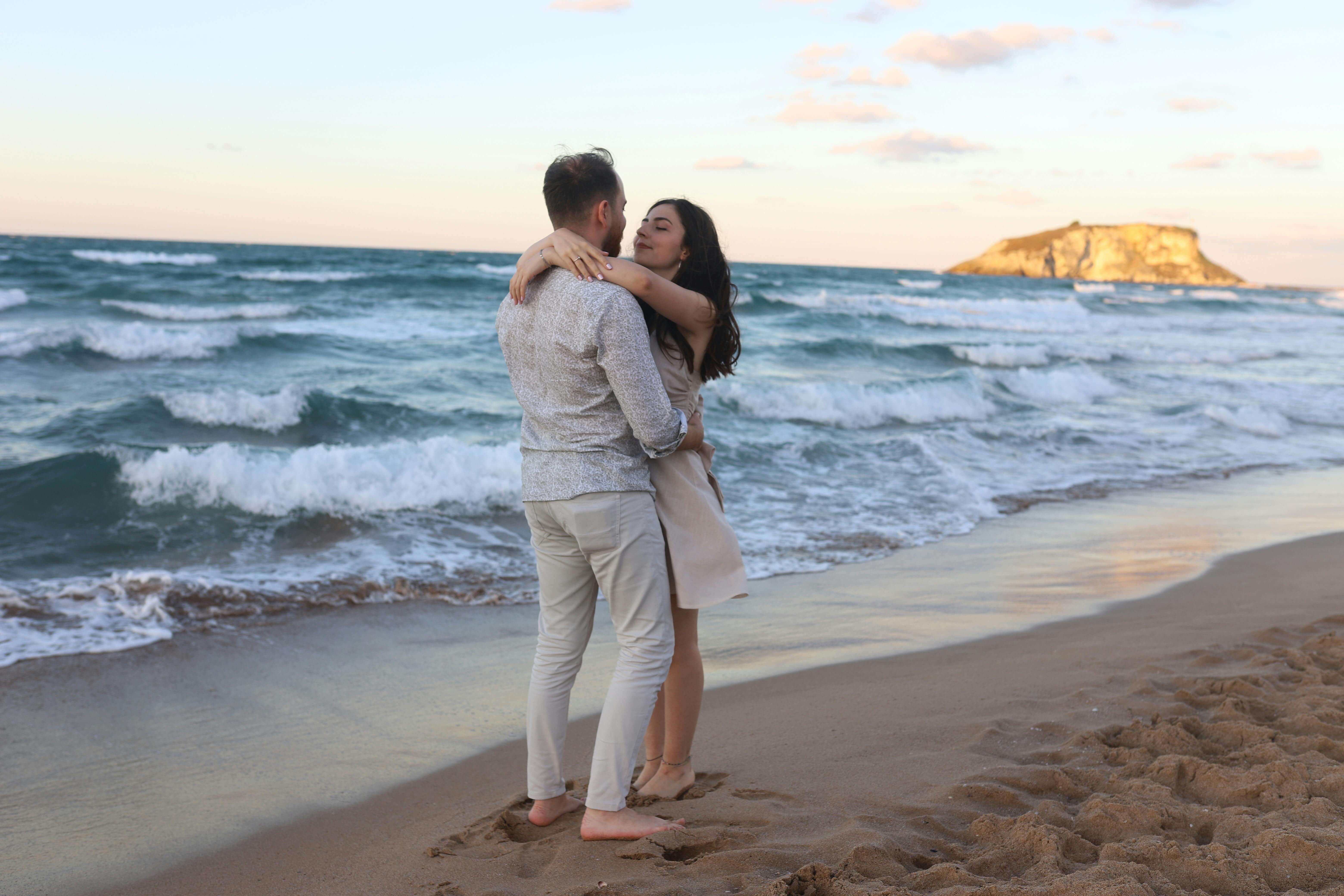 A couple embraces on a sandy beach with waves crashing nearby during a serene sunset.