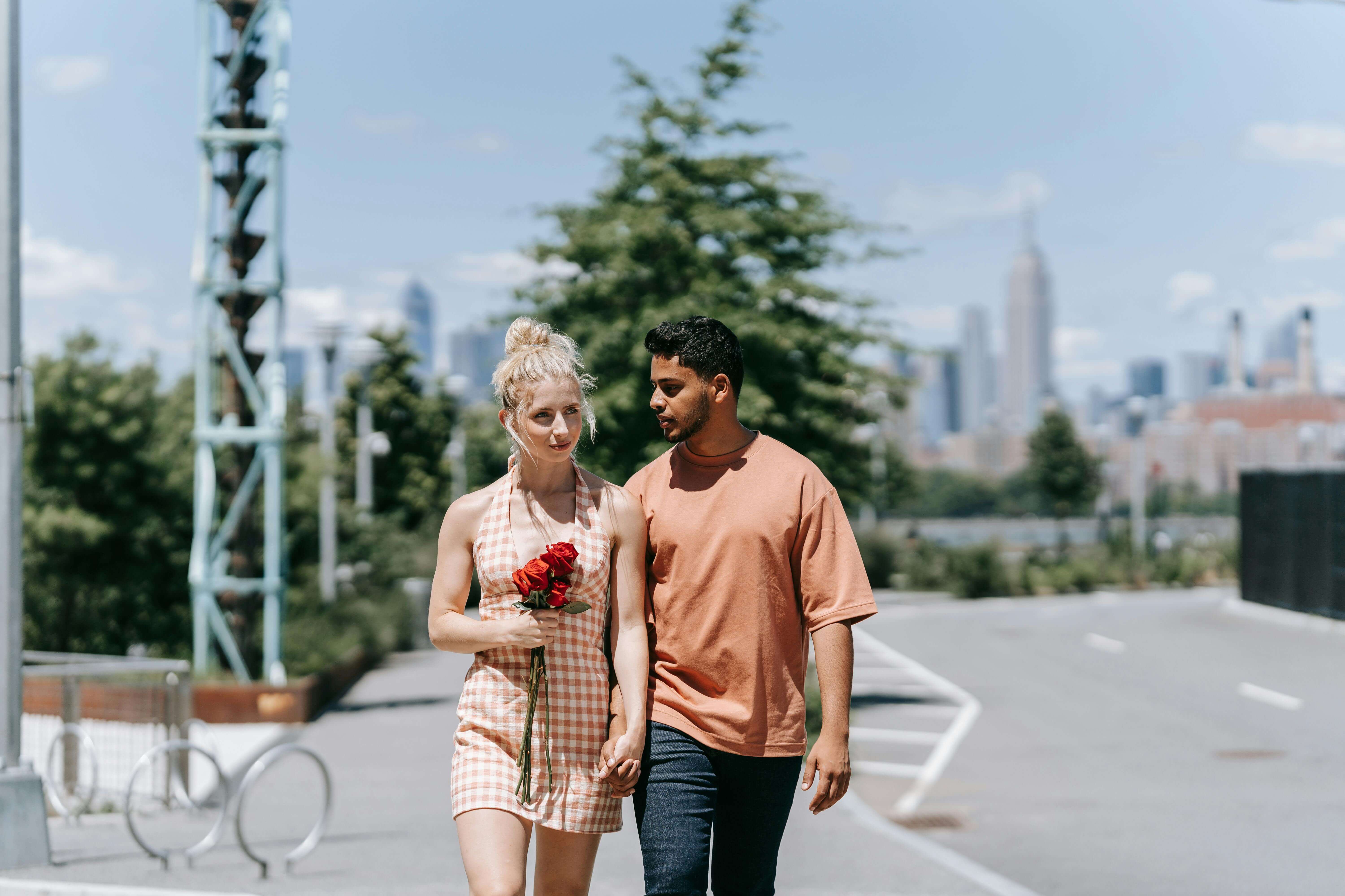 Young couple walking hand in hand outdoors with NYC skyline in the background.
