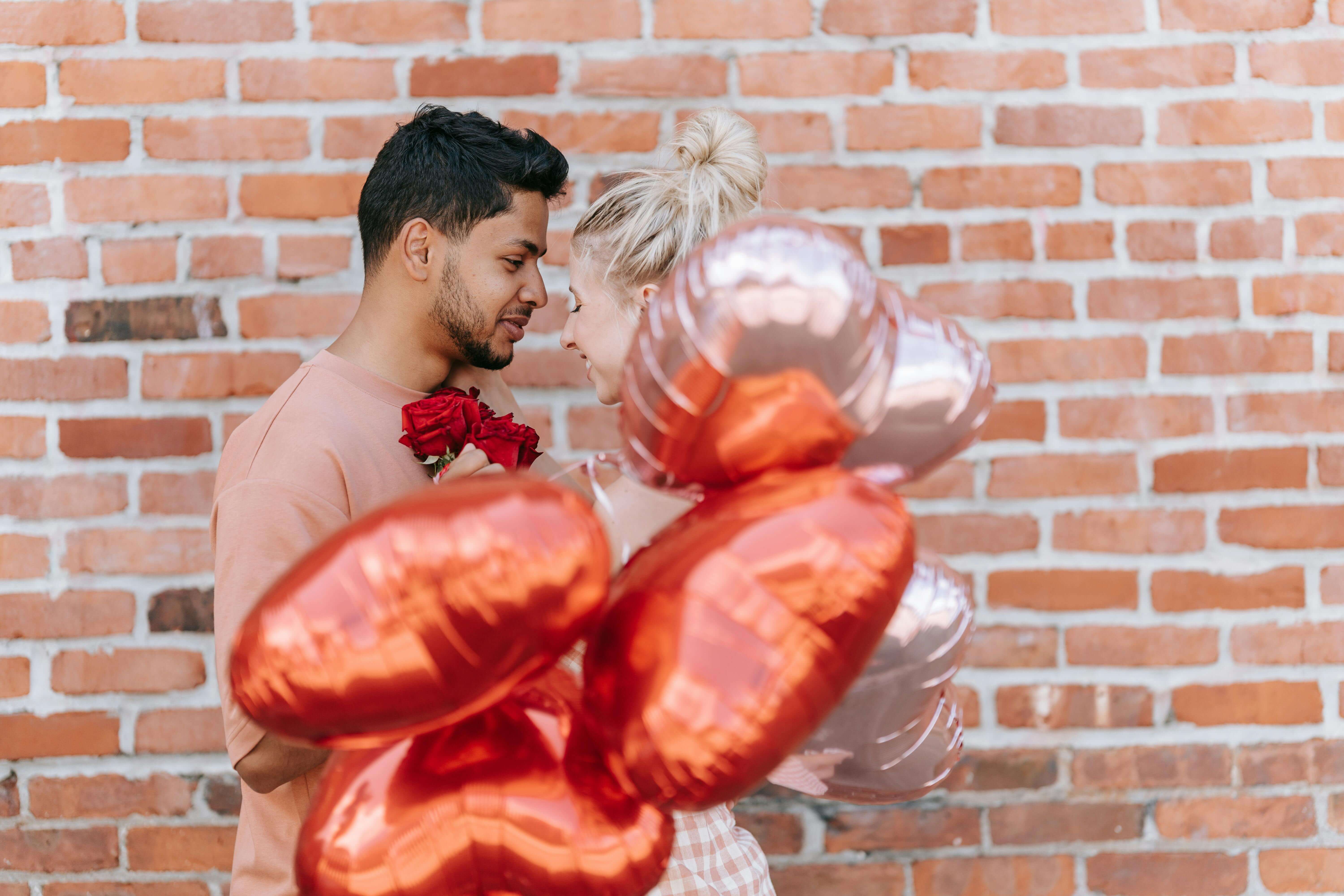 A couple shares an intimate moment with heart-shaped balloons against a brick wall.