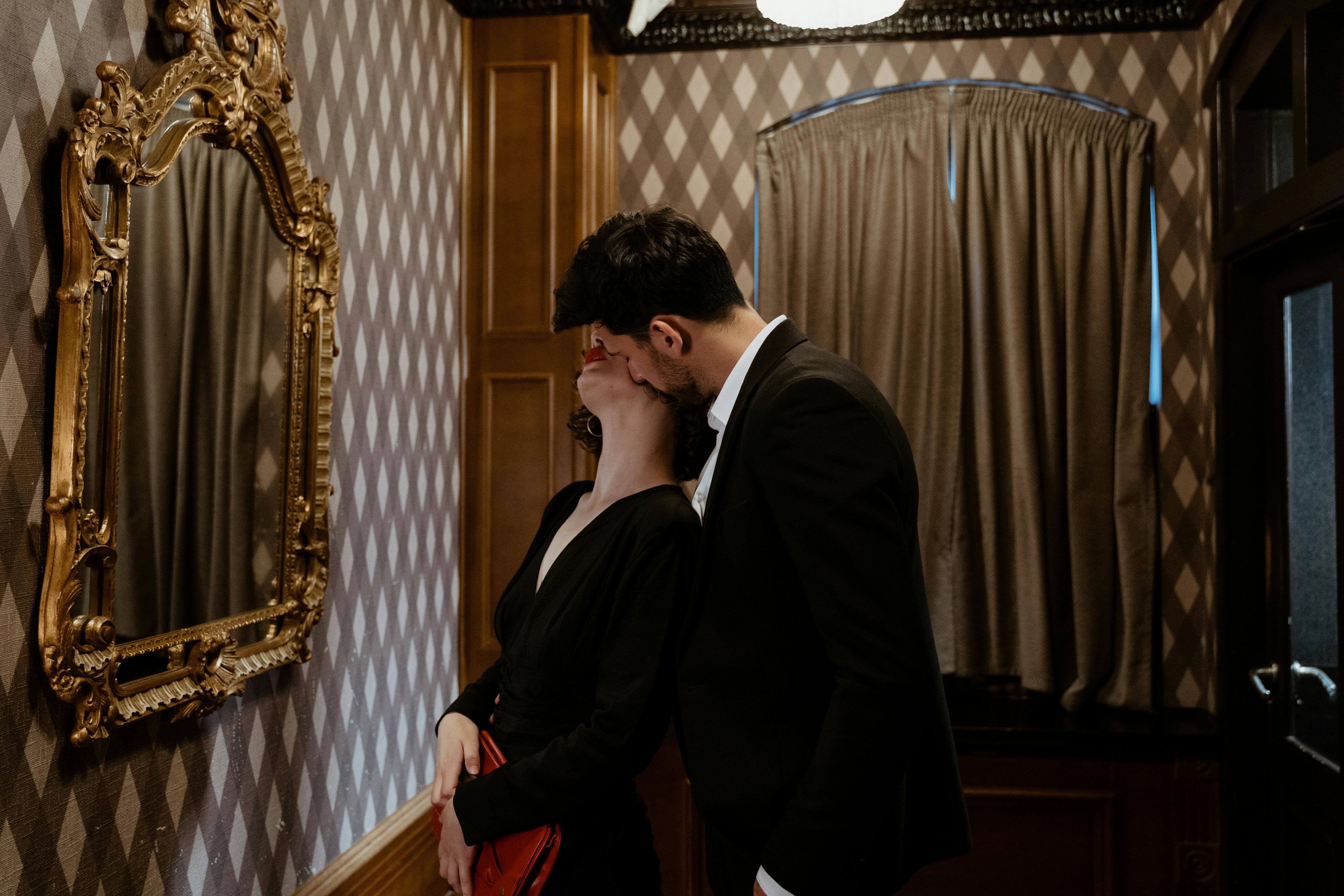 A couple in formal attire shares a tender kiss in an elegant restaurant setting with a vintage mirror.