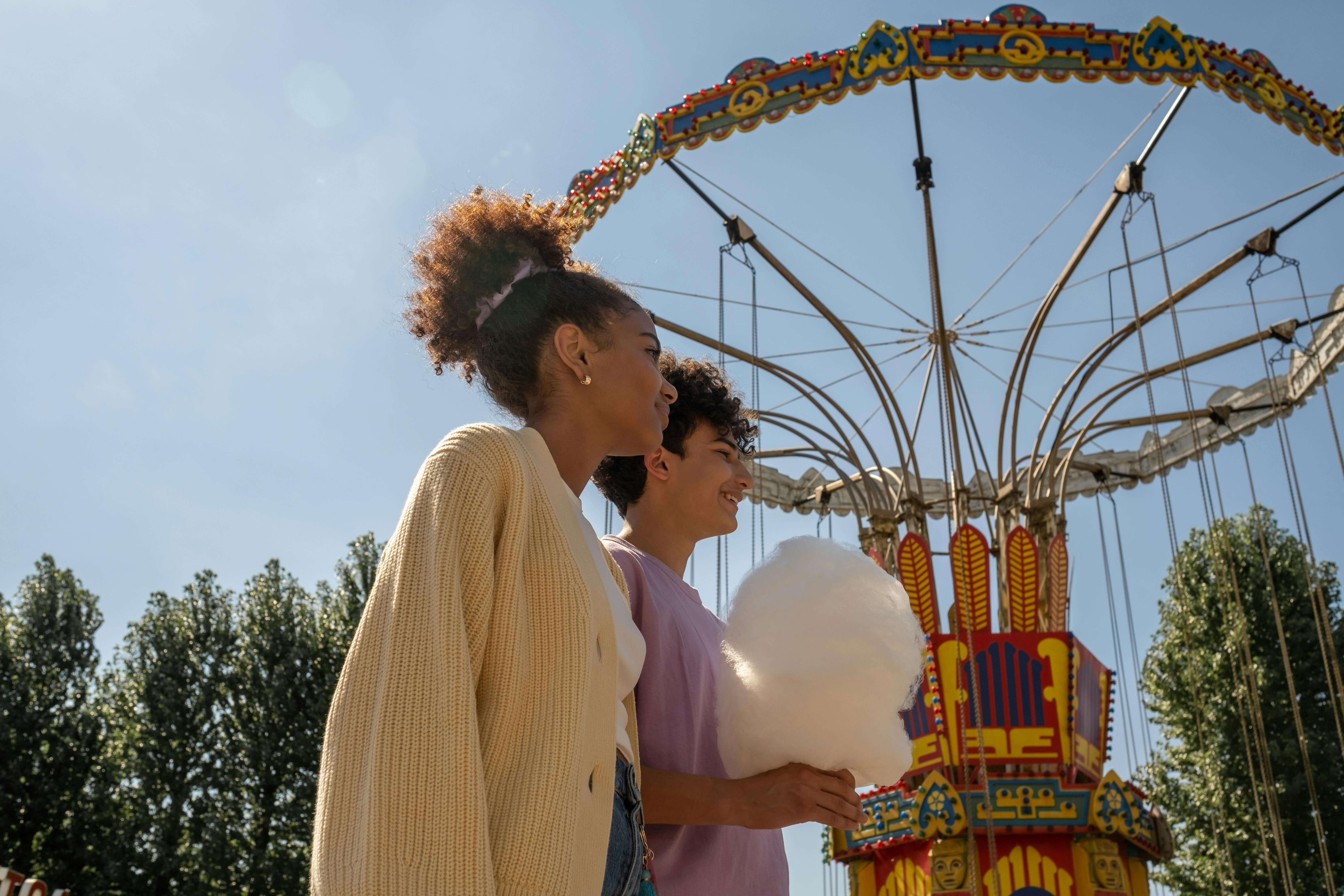 A teenage couple enjoying a sunny day at an amusement park with a carousel and cotton candy.