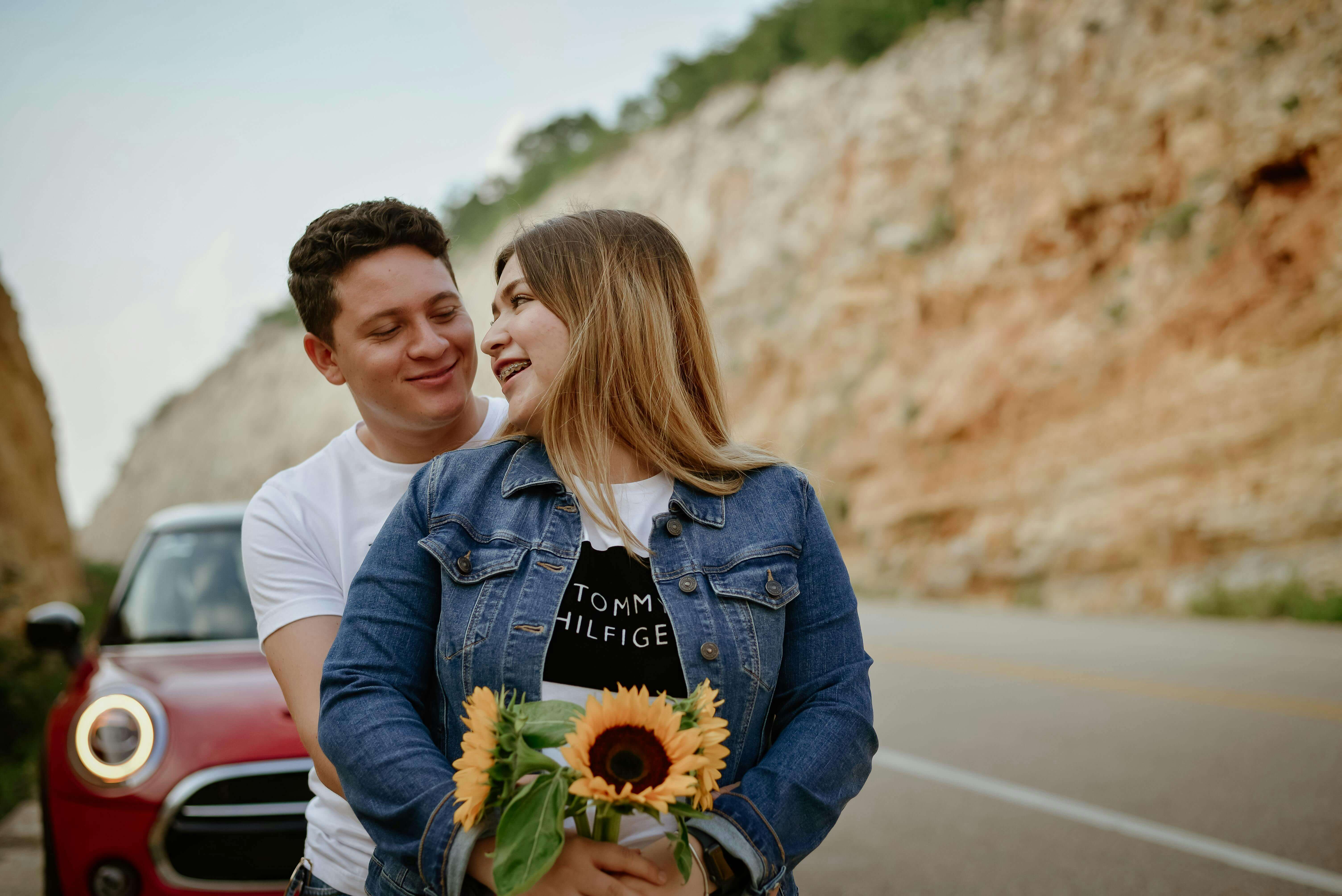 A happy couple shares a joyful moment with sunflowers near a red car on a scenic road.